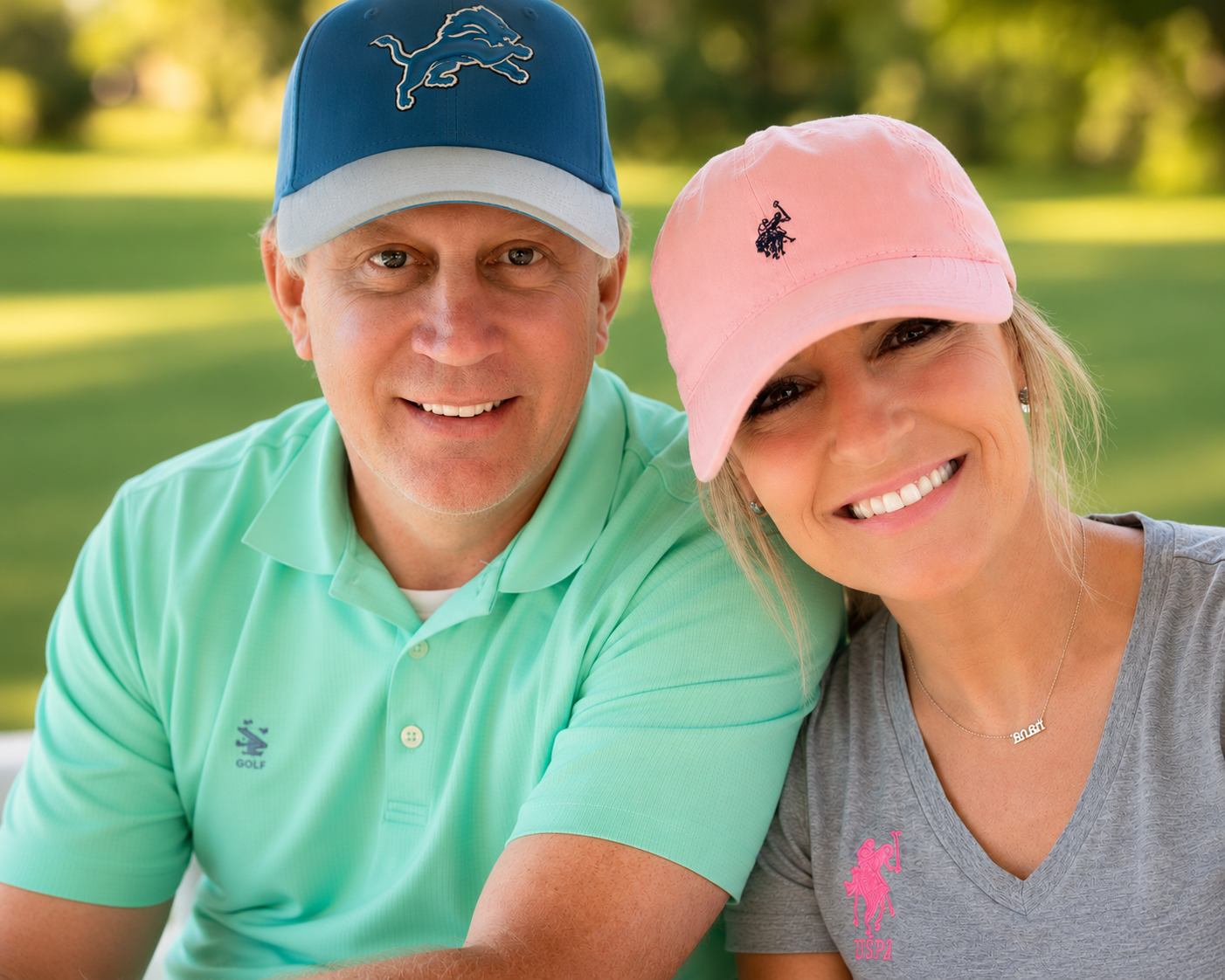 A smiling middle-aged man and woman outdoors in a park, both wearing baseball caps and casual clothing, with a blurred green background.