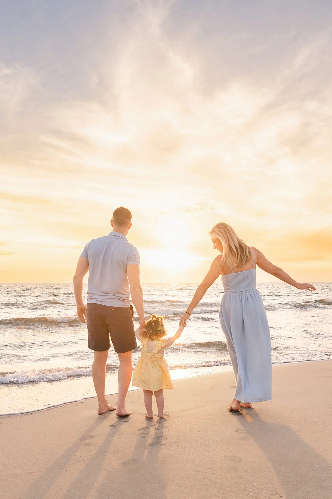 A family consisting of a man, woman, and young girl walking on a beach at sunset. The man and woman are holding the girl's hands, and they are looking at the ocean.