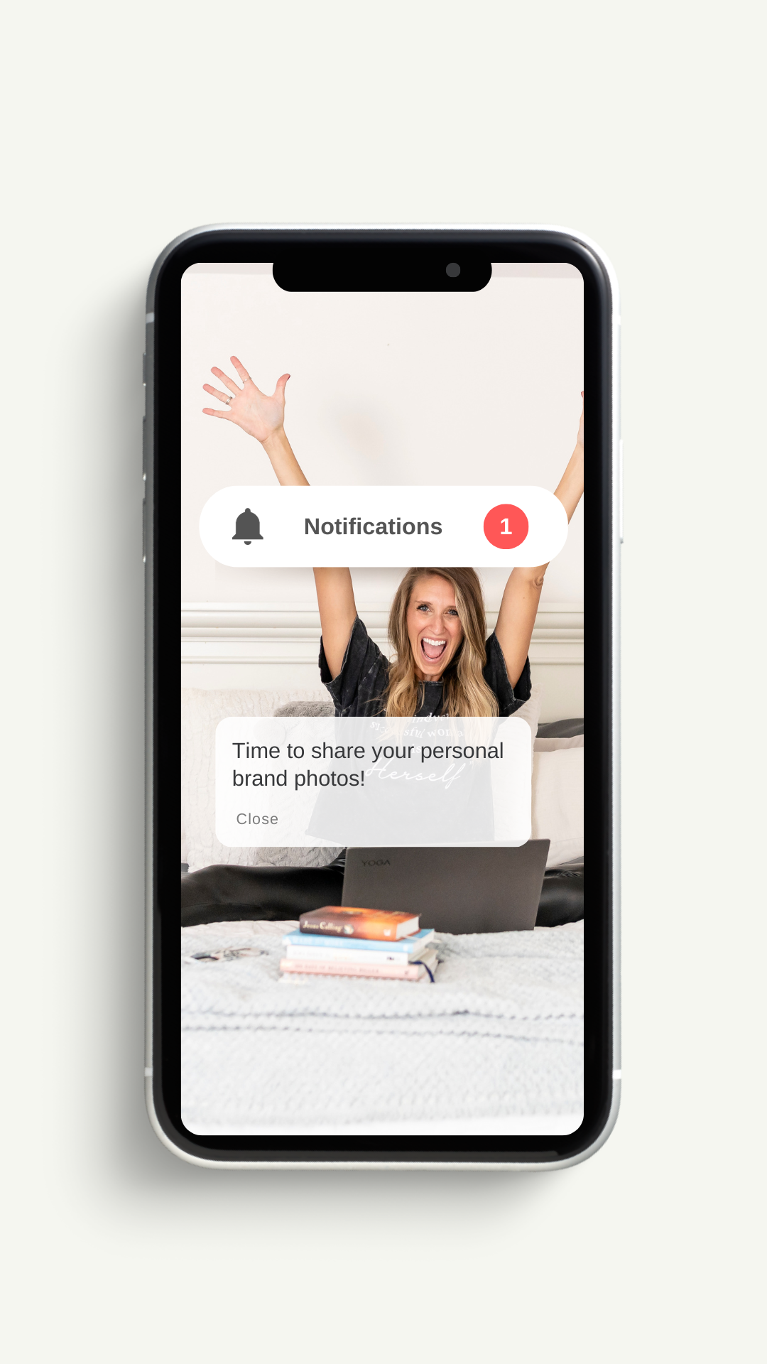 Smartphone screen showing notifications and a woman raising her arms in celebration, sitting on a couch with a laptop and books.