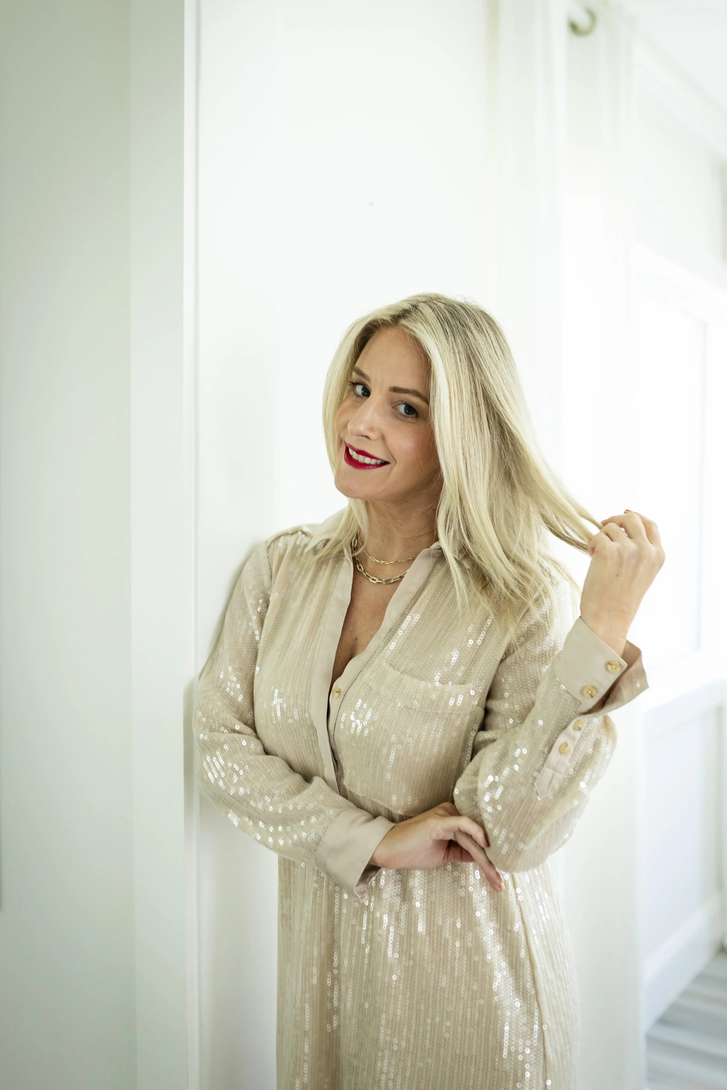 A smiling woman with blonde hair and red lipstick standing against a white wall, wearing a beige sequined blazer and jewelry, with a hand touching her hair.