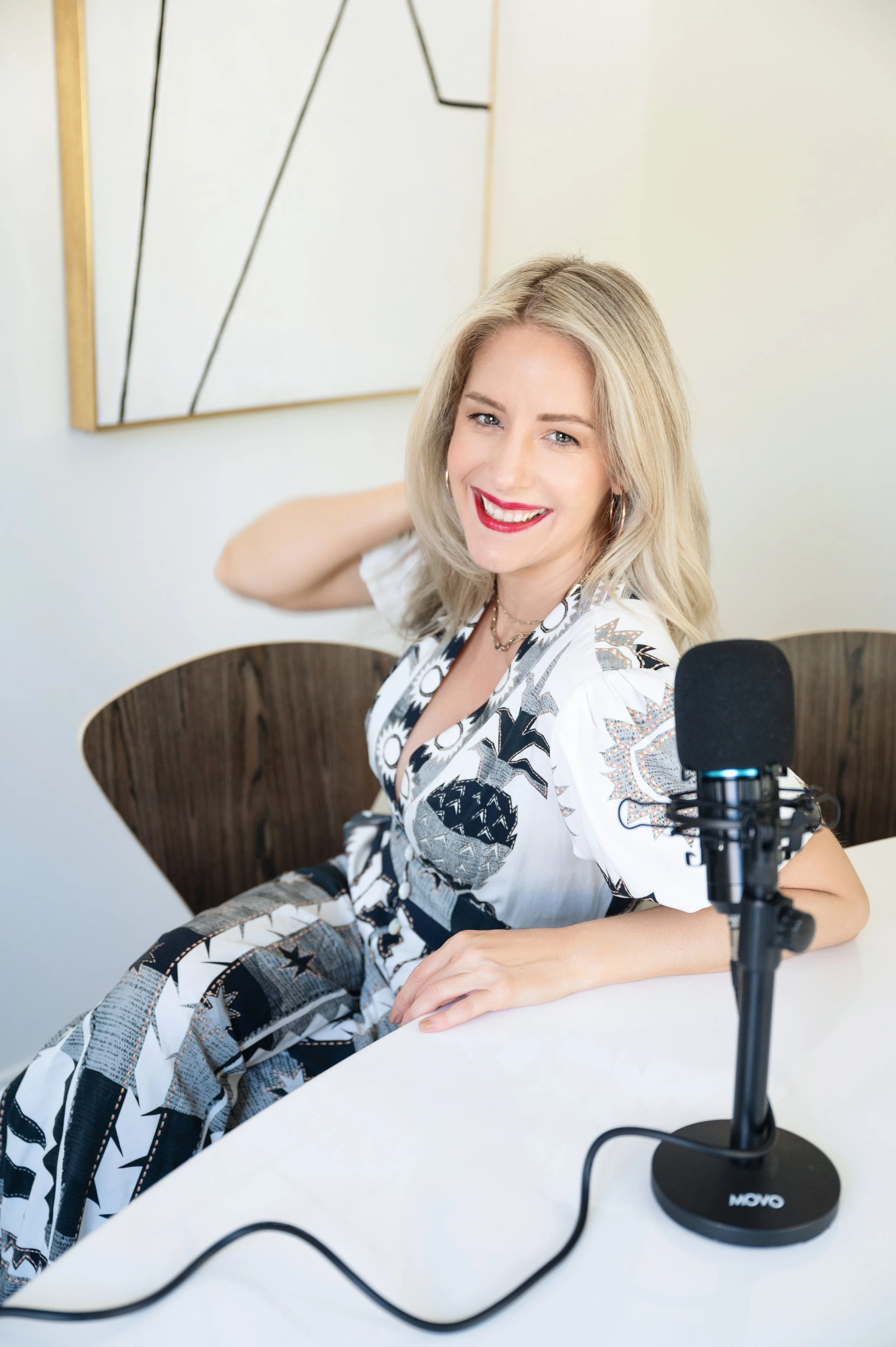 A woman with blonde hair and red lipstick sitting at a white table, smiling, with a professional microphone on a stand in front of her, in a room with beige walls and abstract wall art.