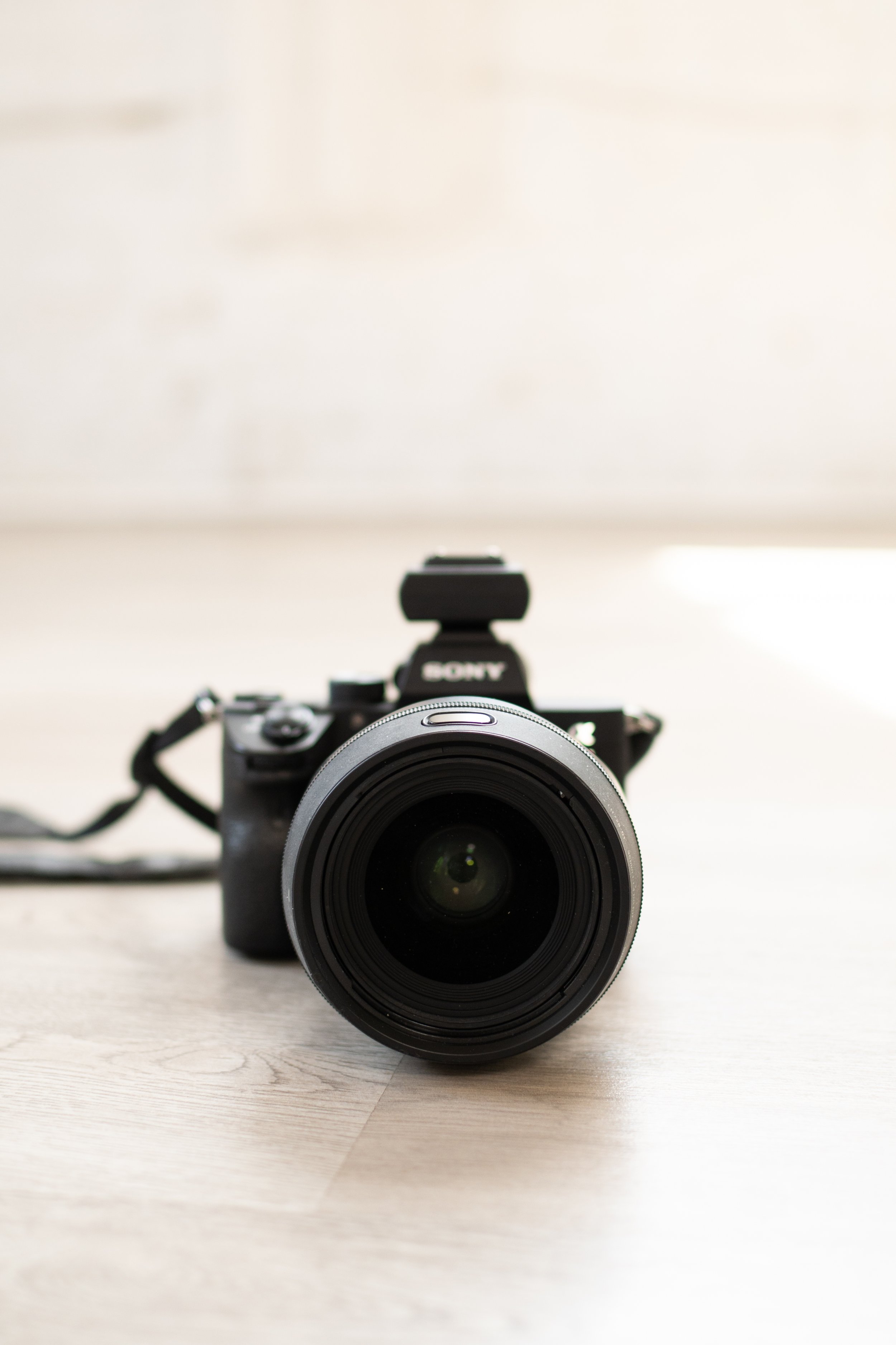 A camera with lens facing forward, placed on a light-colored wooden surface with a blurry, light-colored background.