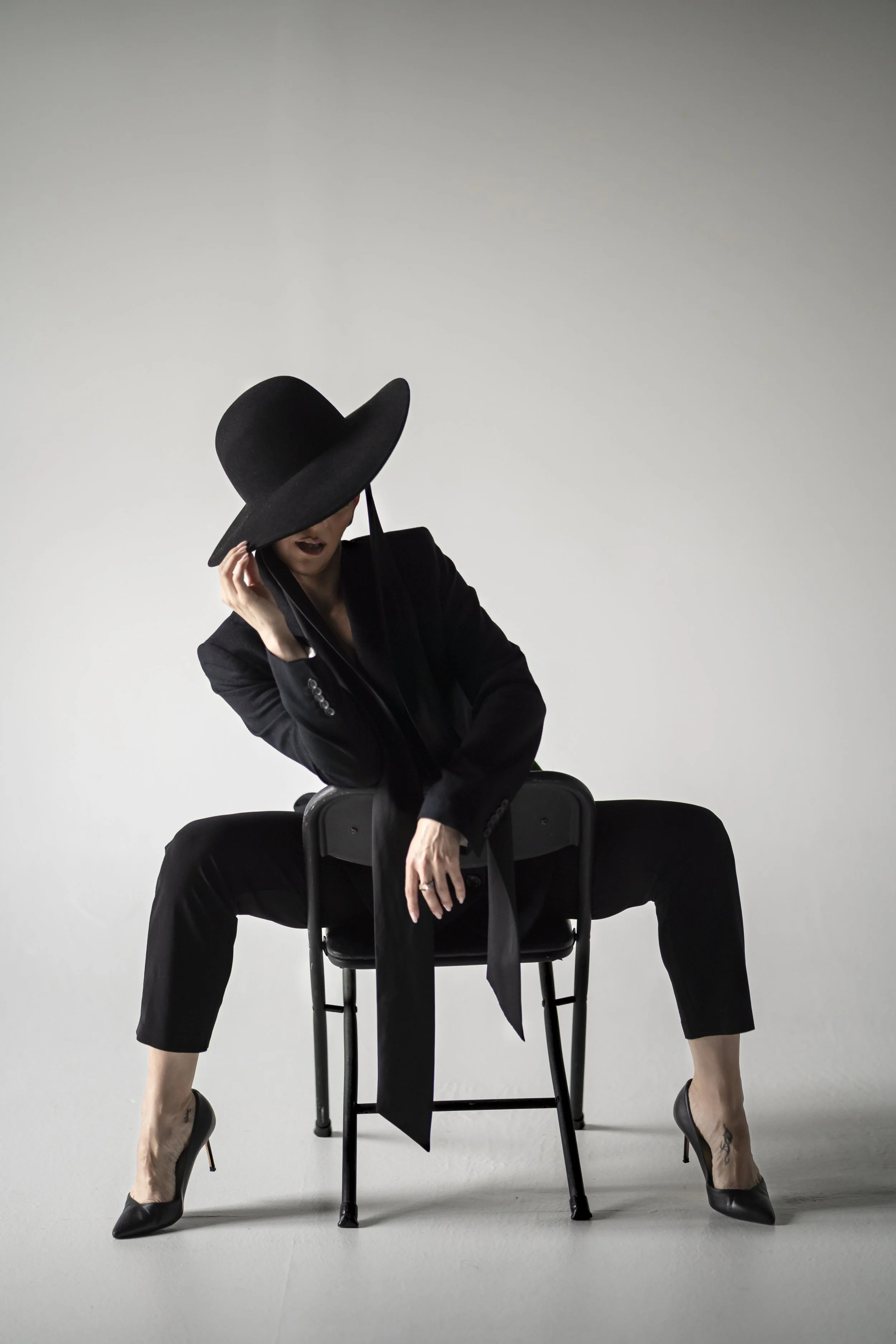 A woman dressed in black, wearing high heels, sitting on a chair. She is holding a large black hat that covers most of her face and head. She is leaning forward with her legs apart and her hands gripping the chair, posing against a plain white background.