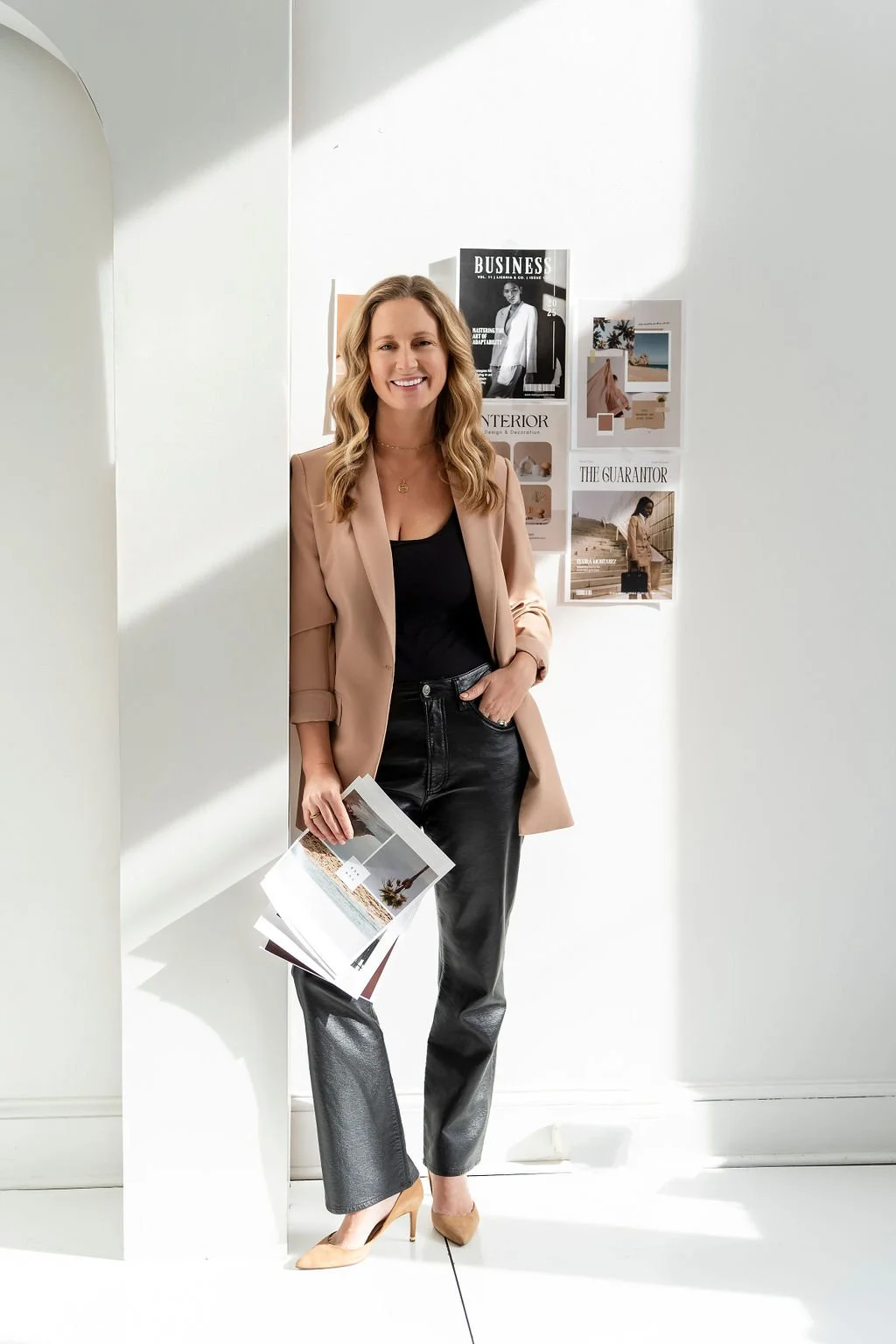 A woman with wavy blonde hair smiling, wearing a beige blazer, black top, black leather pants, and nude heels, standing in a bright room with white walls decorated with magazine pages or posters.