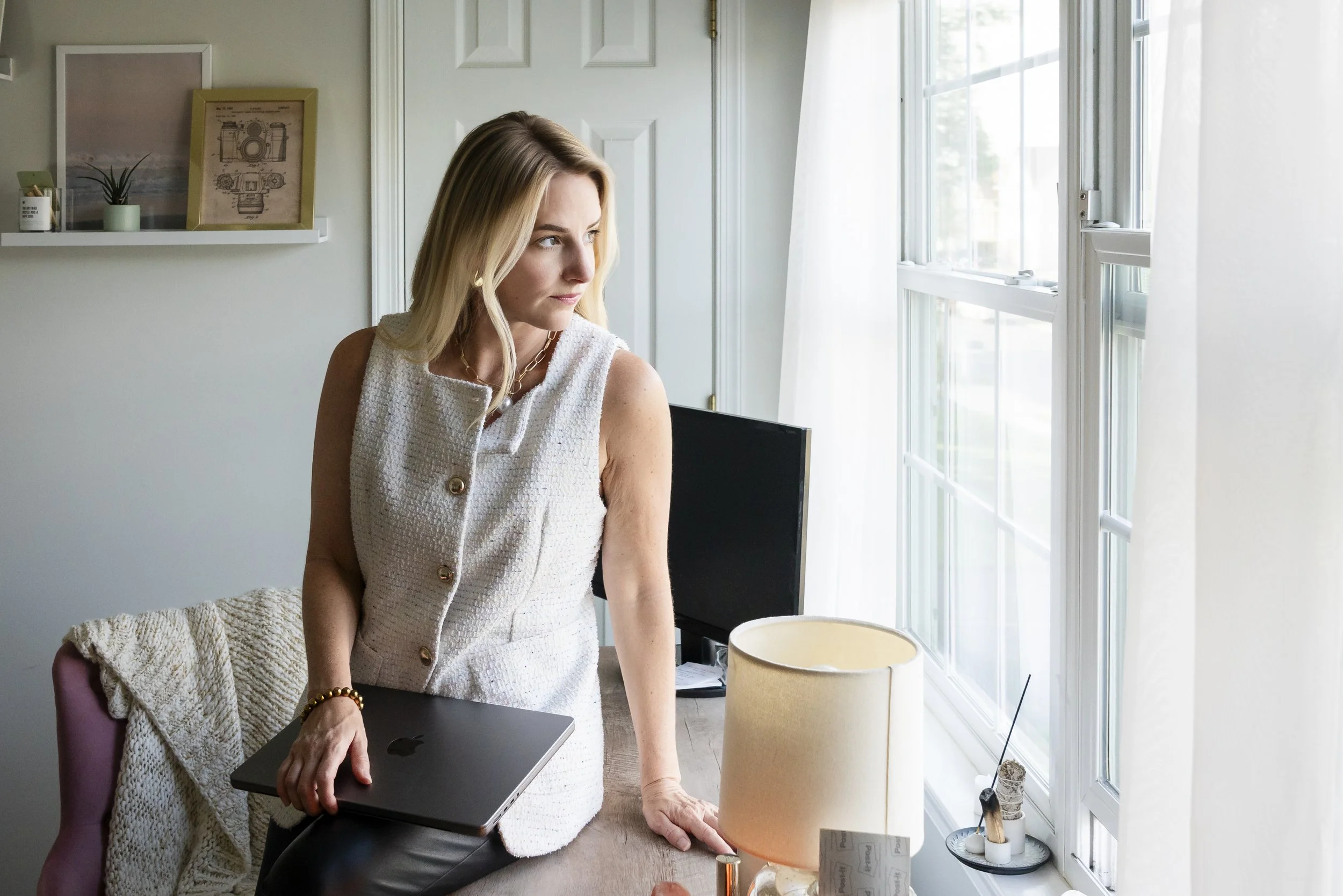 A woman with blonde hair and light skin wearing a white sleeveless dress sitting at a desk, holding a closed MacBook laptop on her lap, in a brightly lit room with large windows and white curtains. There is a beige lamp on the desk, a black chair, and decor with framed pictures and a small plant on a floating shelf on the wall.