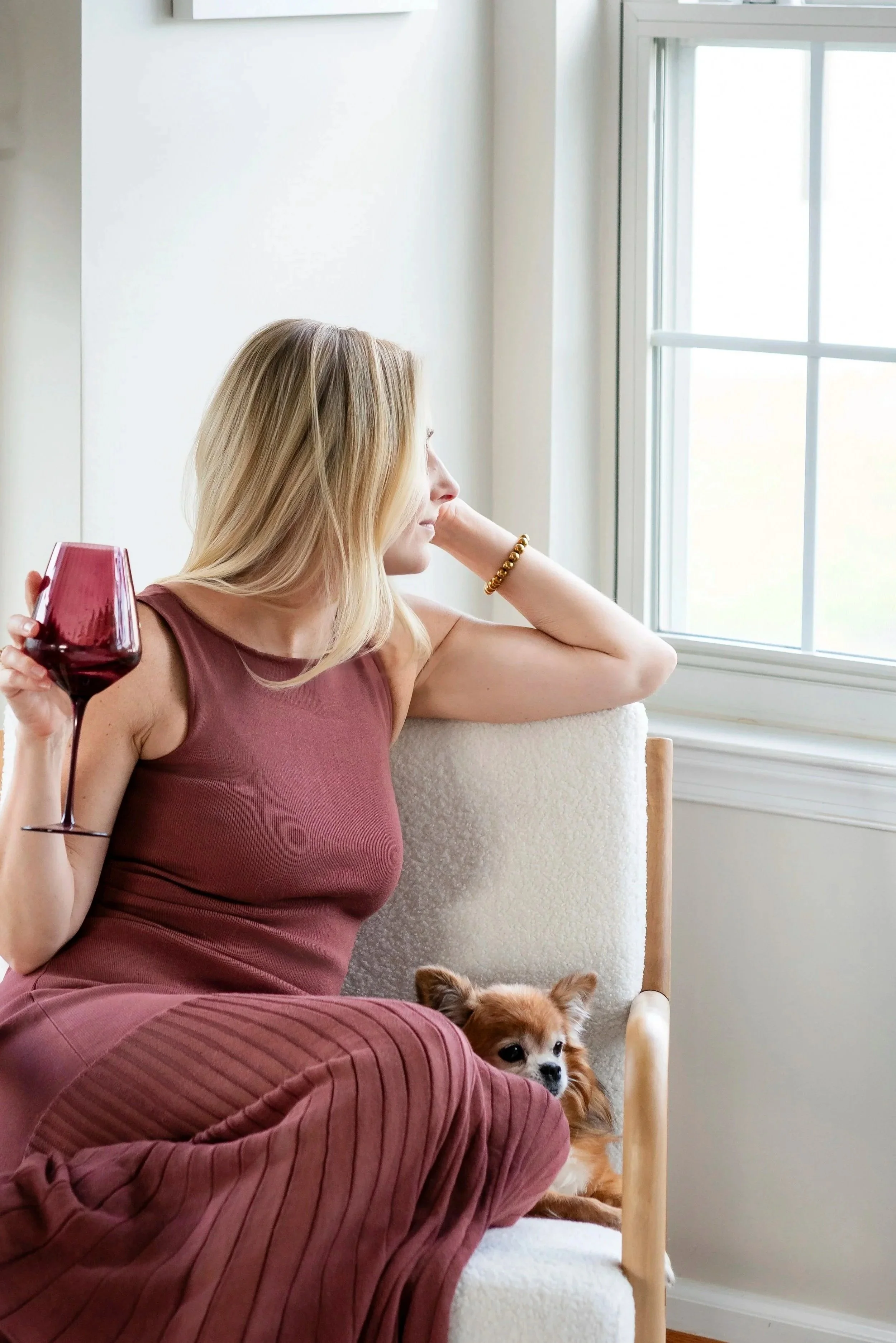 A woman with blonde hair, wearing a maroon top and pants, sits on a chair near a window, holding a glass of red wine. She looks out the window with a contemplative expression. A small brown and white dog sits on her lap, looking towards the camera.