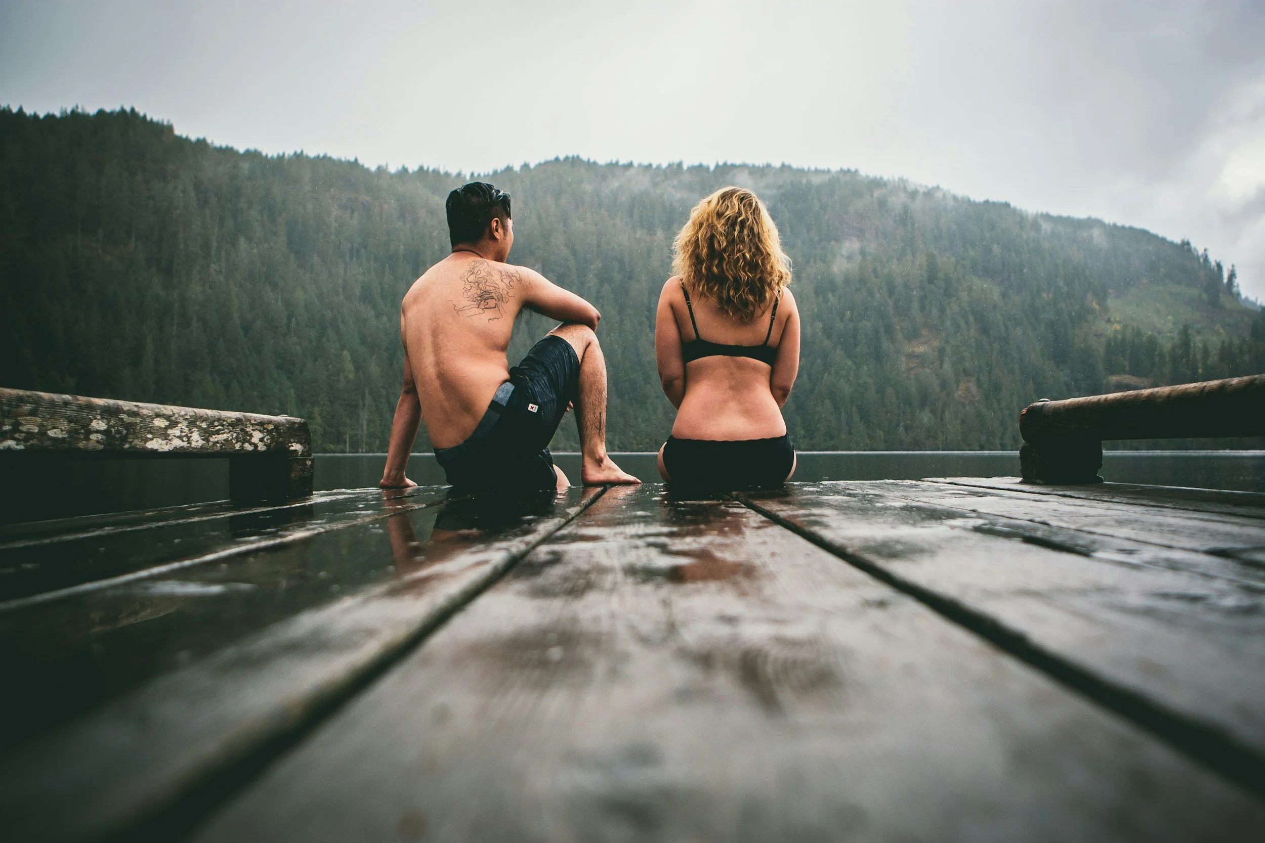 A man with short dark hair and tattoos on his back sits on a wooden dock next to a woman with curly blonde hair, both facing a mountain range covered in trees, with overcast skies in the background.