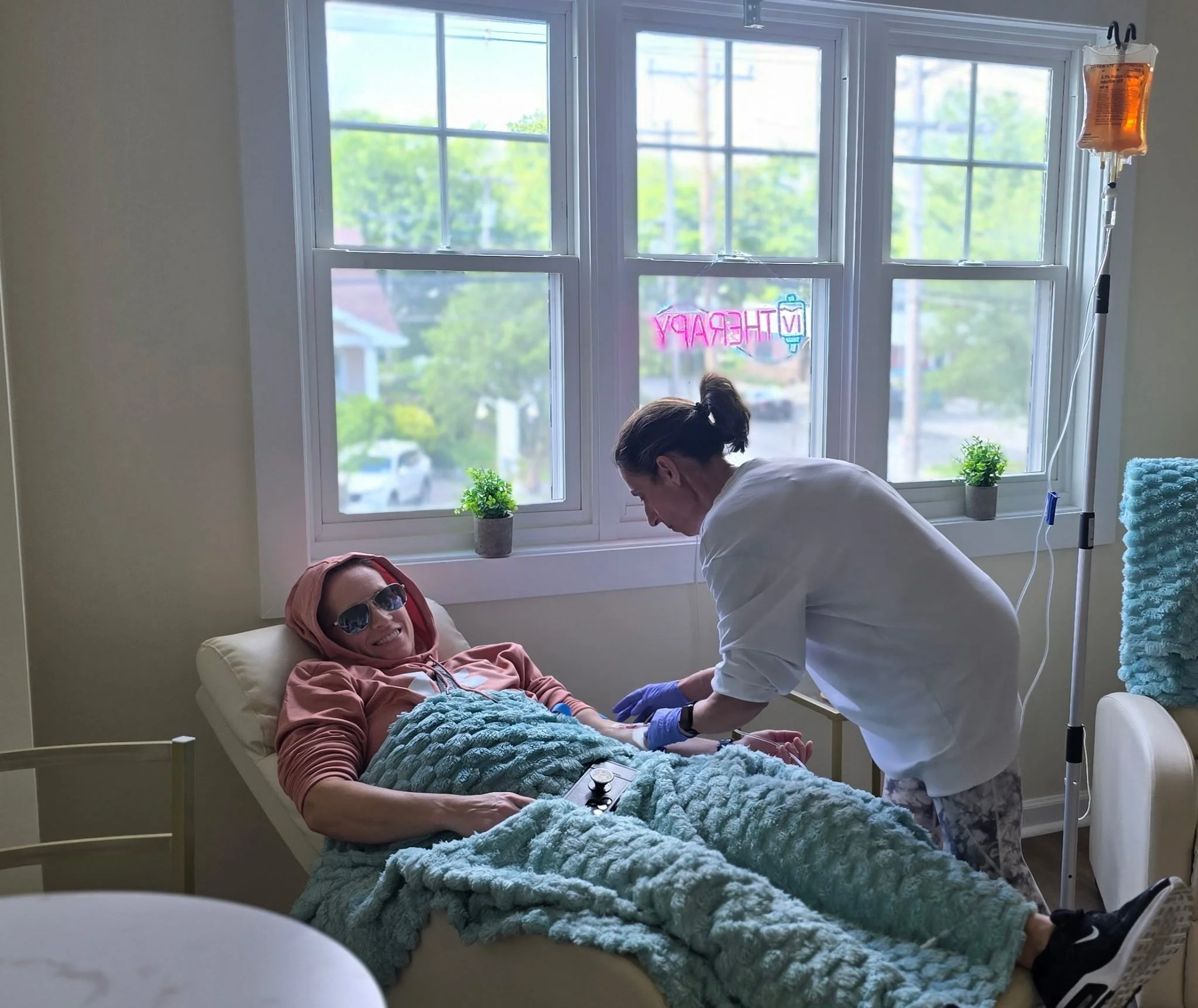 A woman lying in reclining chair bed with headphones, smiling, as a nurse practitioner attends to her in a room with large windows, plants, and a neon sign that says 'IV therapy.