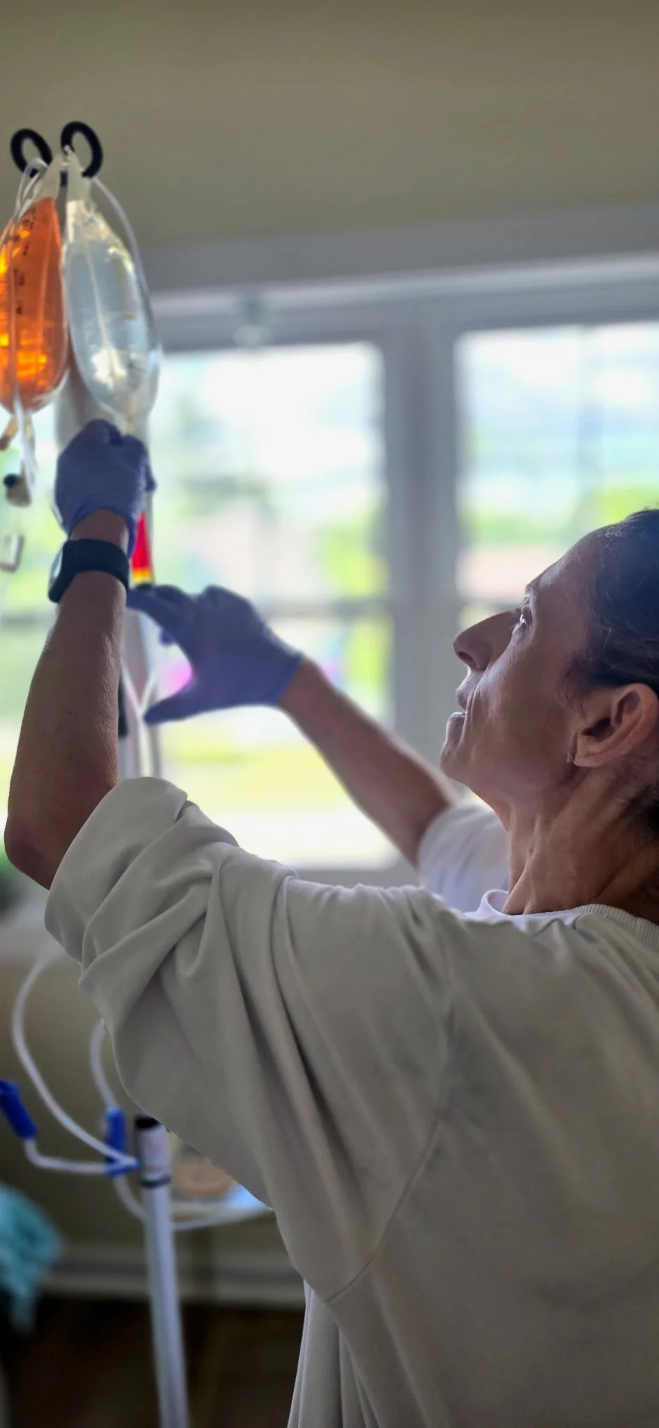 An practitioner wearing a white lab coat and purple gloves reaching up to adjust IV bags hanging from a medical stand in a hospital room with windows and green scenery outside.