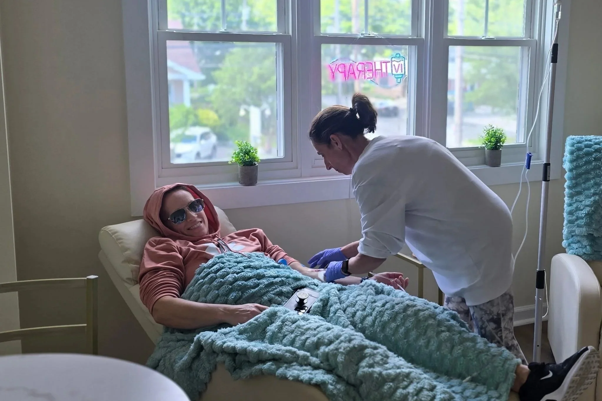 A woman lying in a hospital bed with sunglasses and a hoodie, smiling. A healthcare worker in white scrubs and purple gloves attending to her. The room has a large window with a neon sign saying 'Emergency' behind them, and two small plants on the windowsill.