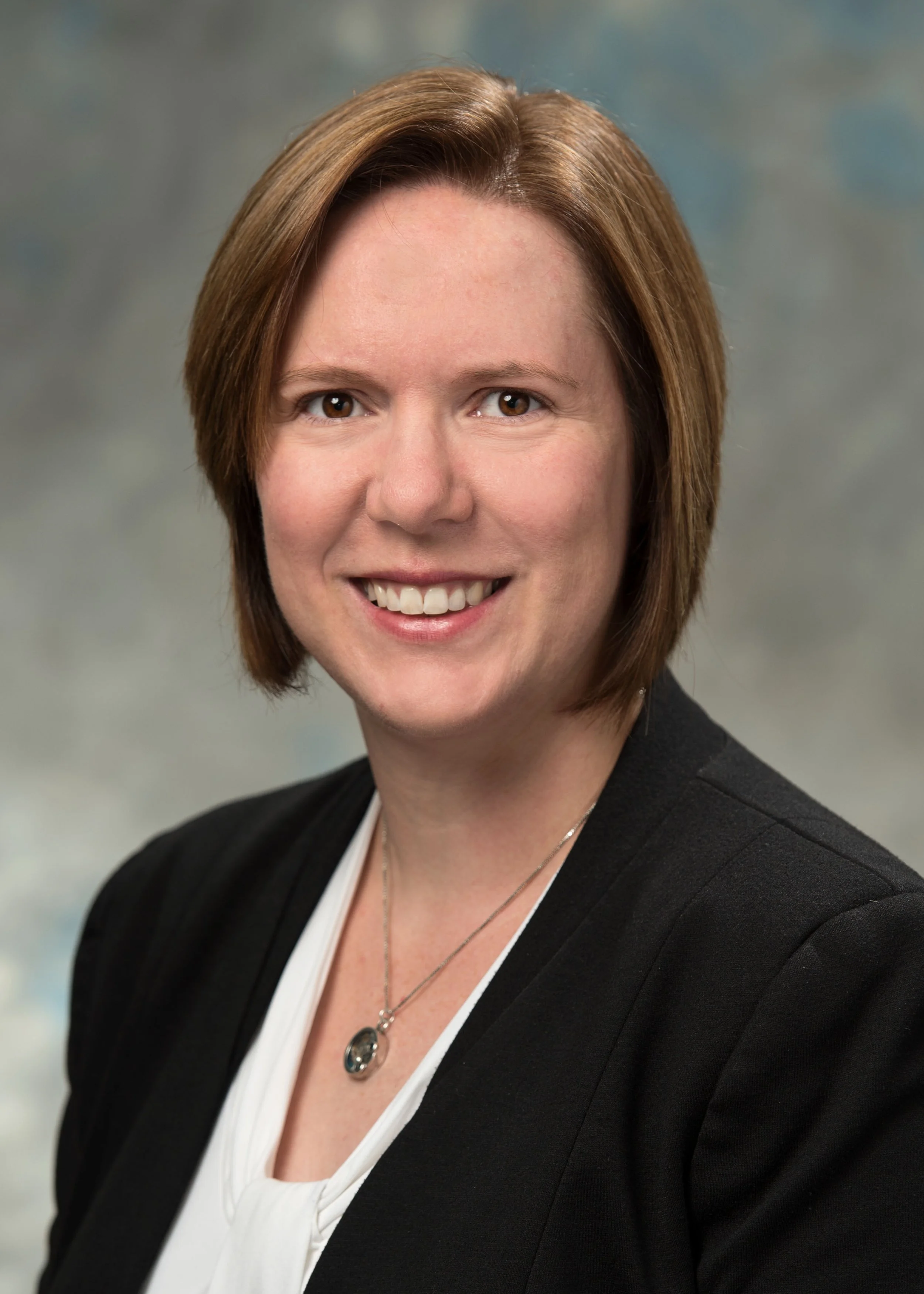 Headshot of a woman with brown hair, smiling, wearing a black blazer over a white top, with a necklace, against a blurred background.