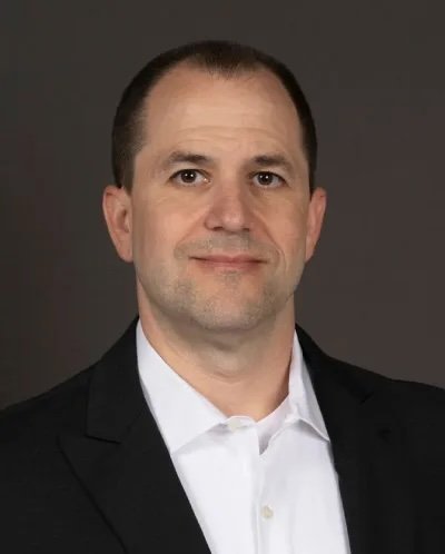 A professional headshot of a man with short dark hair, wearing a black suit jacket and white shirt, against a gray background.