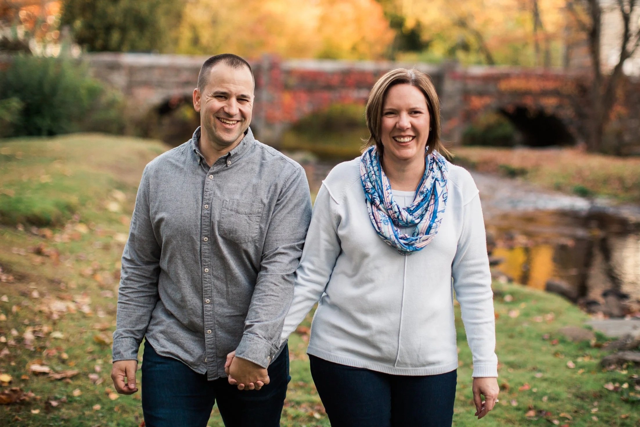 A man and woman walking outdoors in a park during fall, holding hands, smiling, with colorful autumn trees and a bridge in the background.