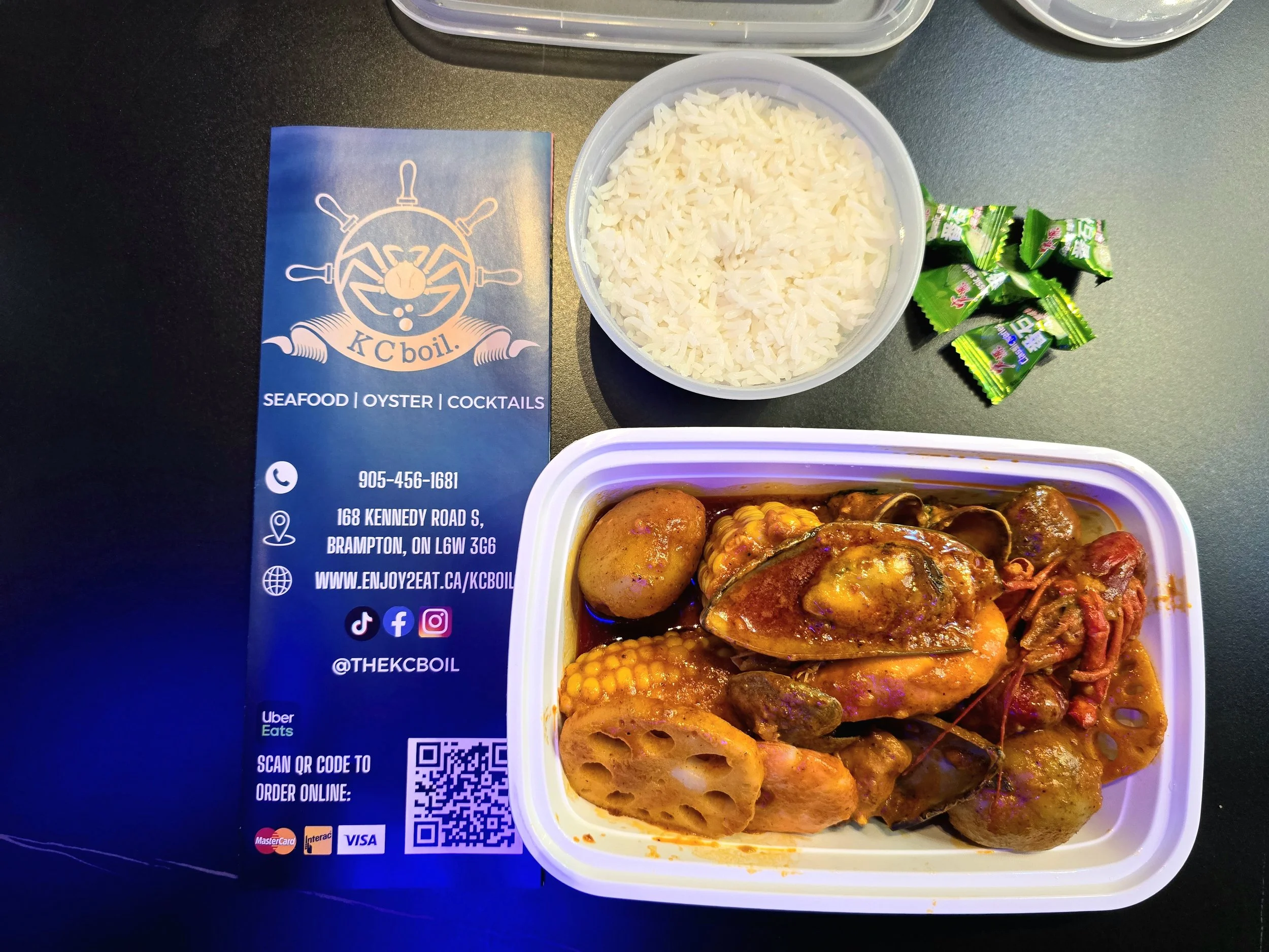 A container of cooked white rice, a few fortune cookies, and a portion of assorted seafood and meat in sauce, with a tray and business card for KC Boil seafood restaurant nearby.