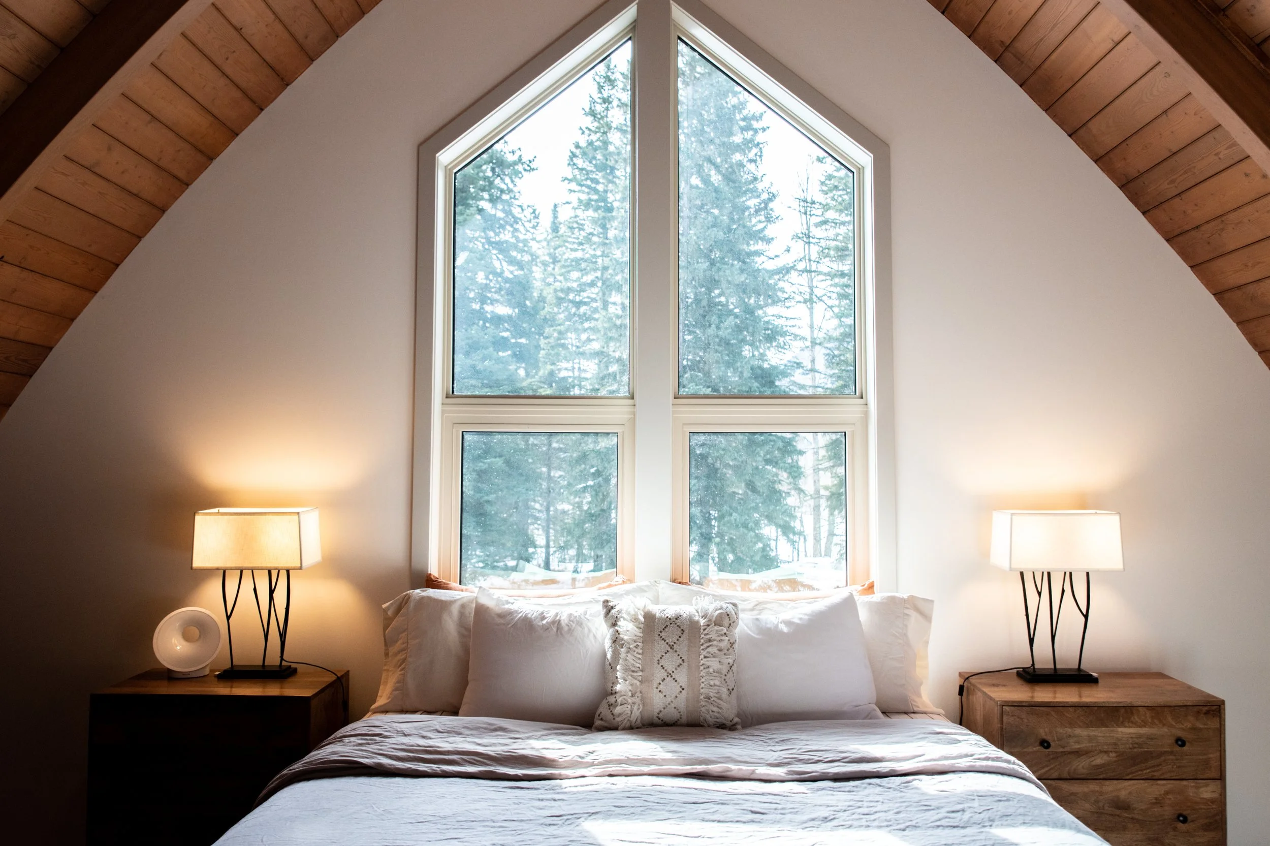 A bedroom with a large pyramid-shaped window showing a snowy outdoor landscape with tall trees, with a bed in front and two matching bedside tables with lamps.