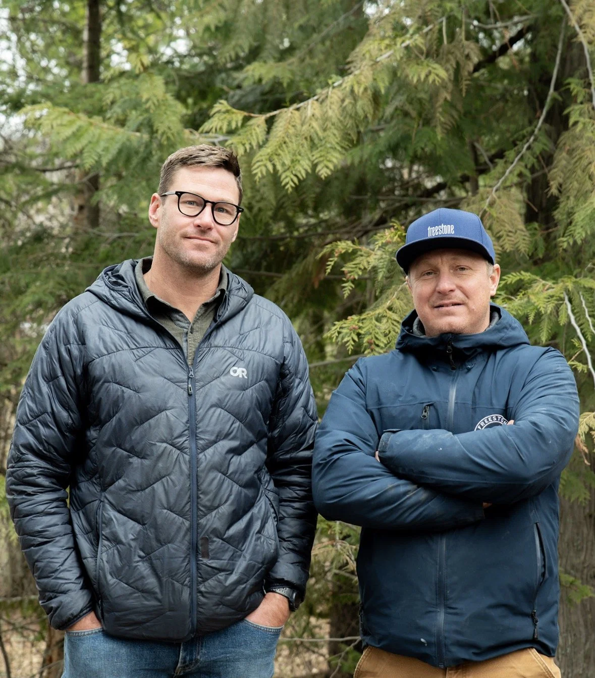 Two men standing outdoors in front of a tree. The man on the left has short brown hair, glasses, and is wearing a black quilted jacket and blue jeans. The man on the right has short hair, is wearing a blue cap and jacket, and has his arms crossed.