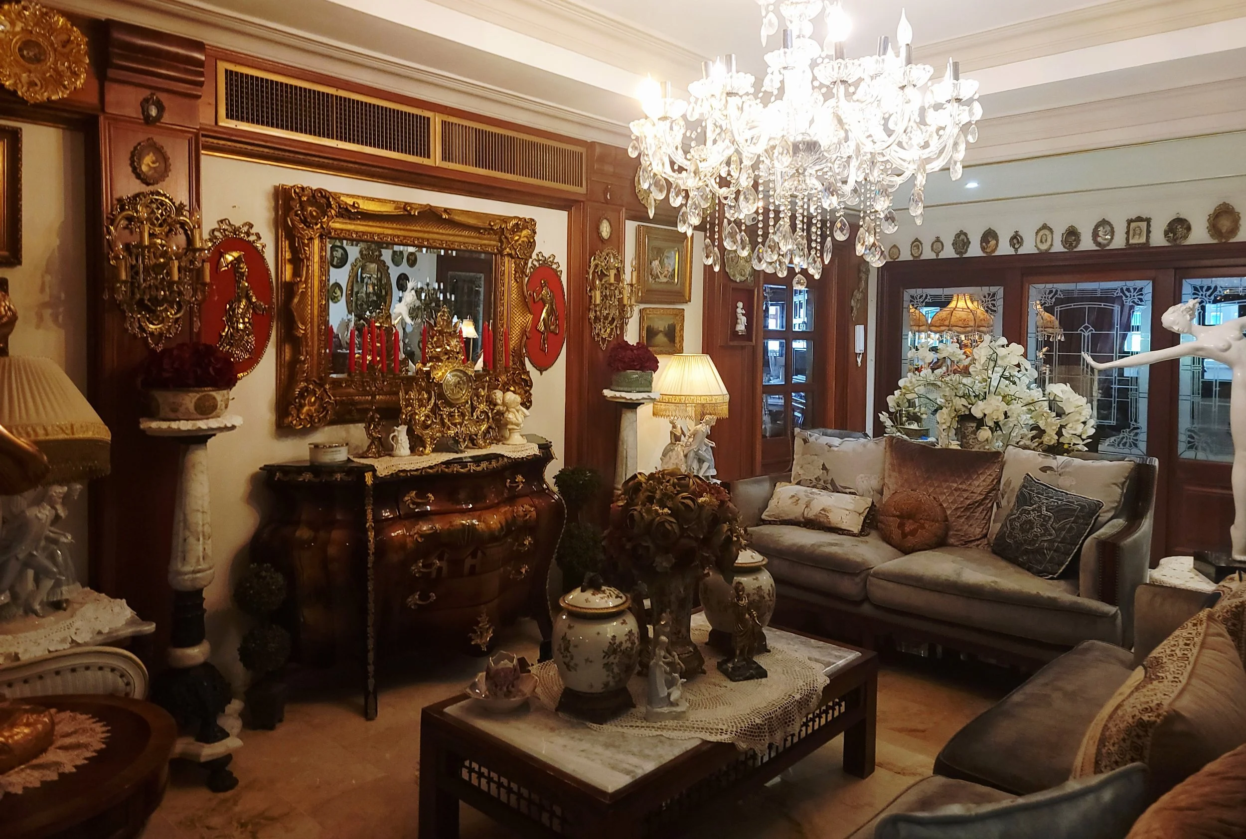 A luxurious living room with ornate gold-framed mirrors, vintage decor, a crystal chandelier, upholstered sofas, and decorative vases.