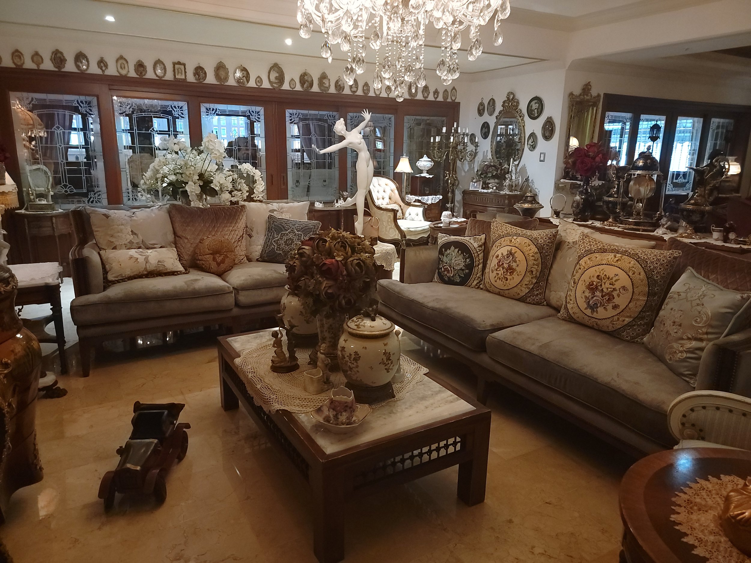 A living room decorated with antique and vintage furniture, including a large sofa with multiple pillows, a wooden coffee table with decorative ceramics, and floral arrangements. The room has a chandelier hanging from the ceiling, and walls adorned with decorative plates and mirrors. Large windows with intricate patterns on the glass allow natural light to enter.