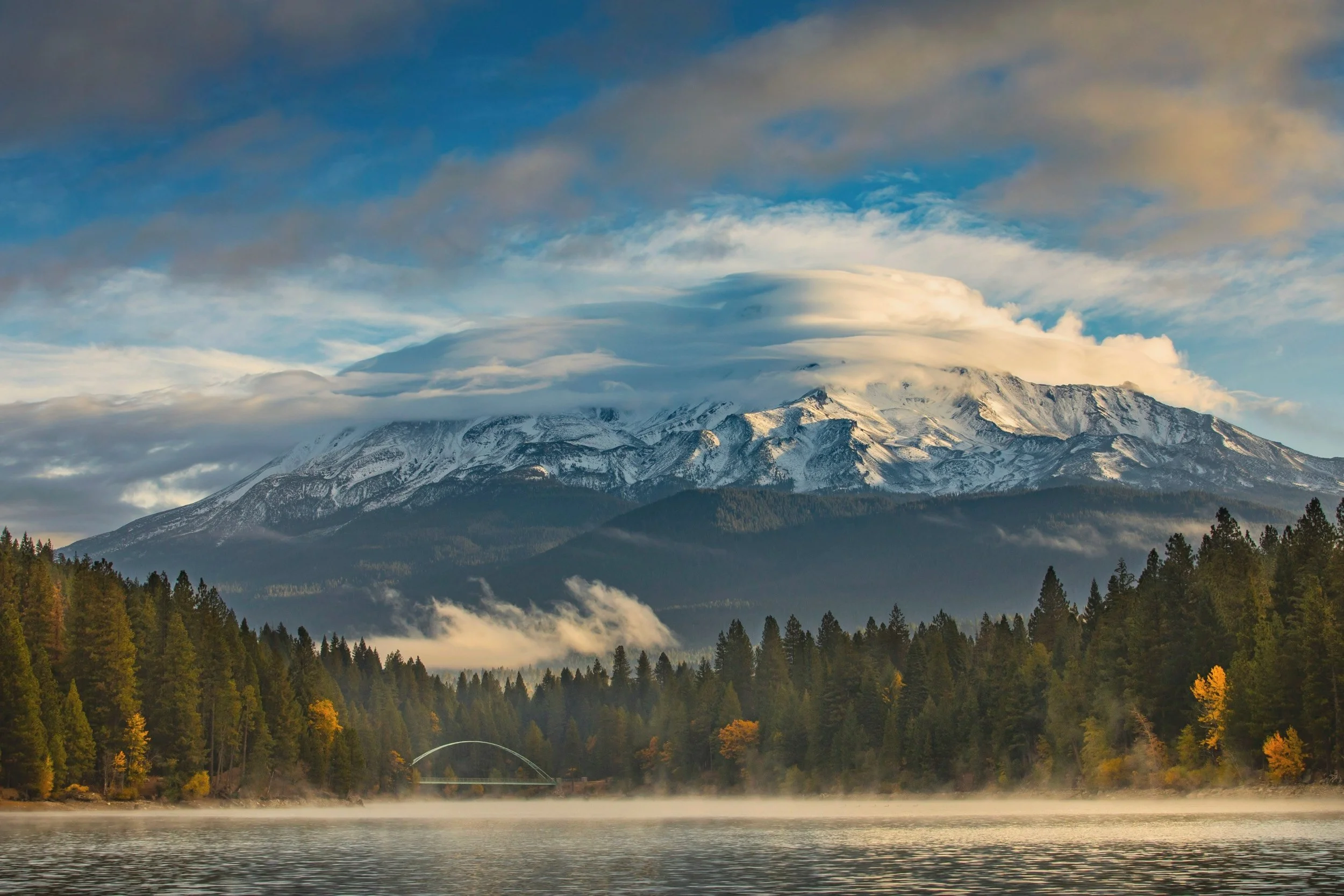 Snow-capped mountain behind a forest with green and yellow trees, reflecting in a calm river, with clouds and a blue sky overhead.