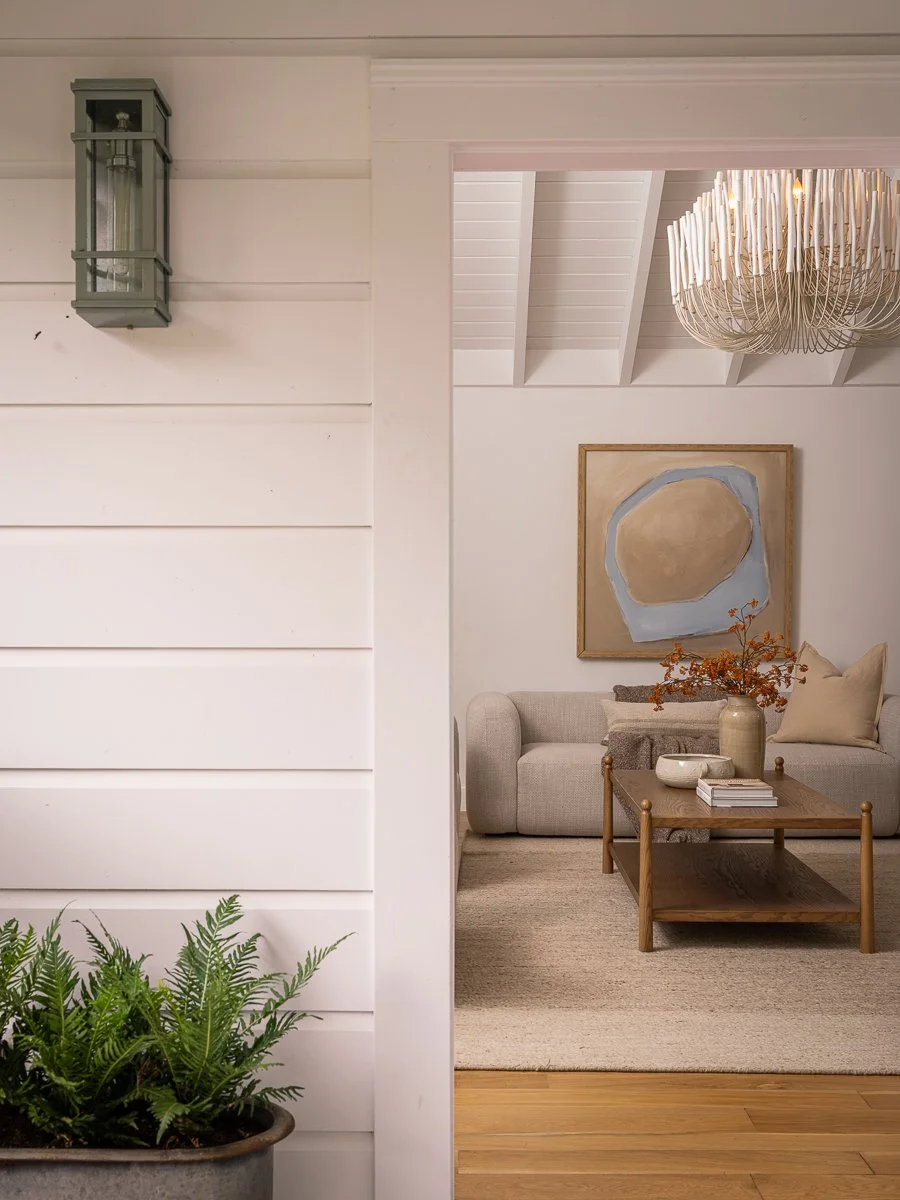 Living room with beige sofa, wooden coffee table, abstract art painting, and large white chandelier. Part of the entryway includes a green potted fern and white shiplap walls.