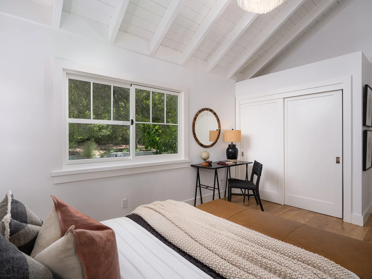A bedroom with a sloped white ceiling, a large window showing greenery outside, a bed with multiple pillows, a wooden bench, and a small black desk with a black chair, a black table lamp, a round mirror, and decorative objects.