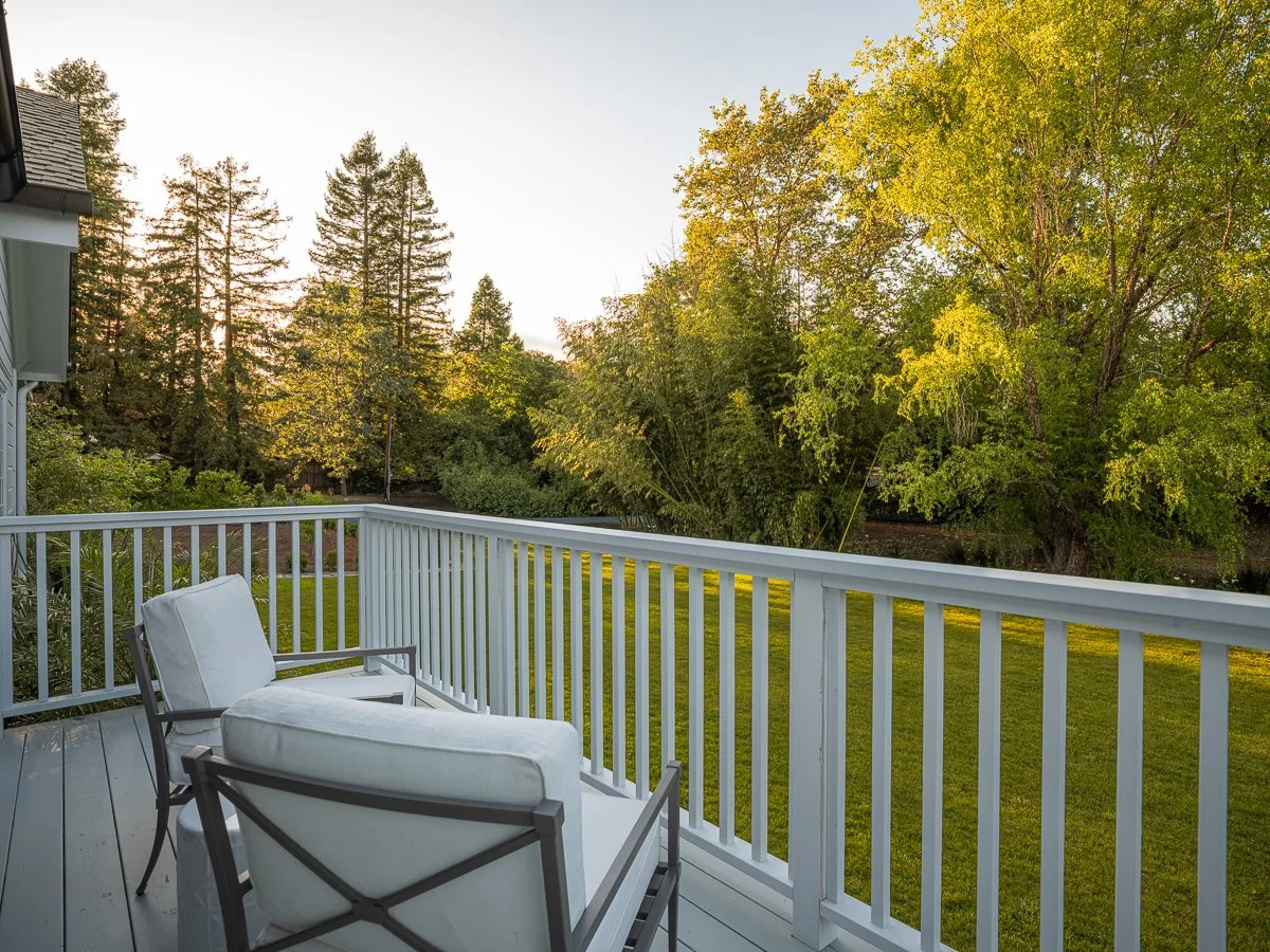 View of a backyard with a wooden deck, white chairs with cushions, green grass, and tall trees with green leaves in the distance during sunset.