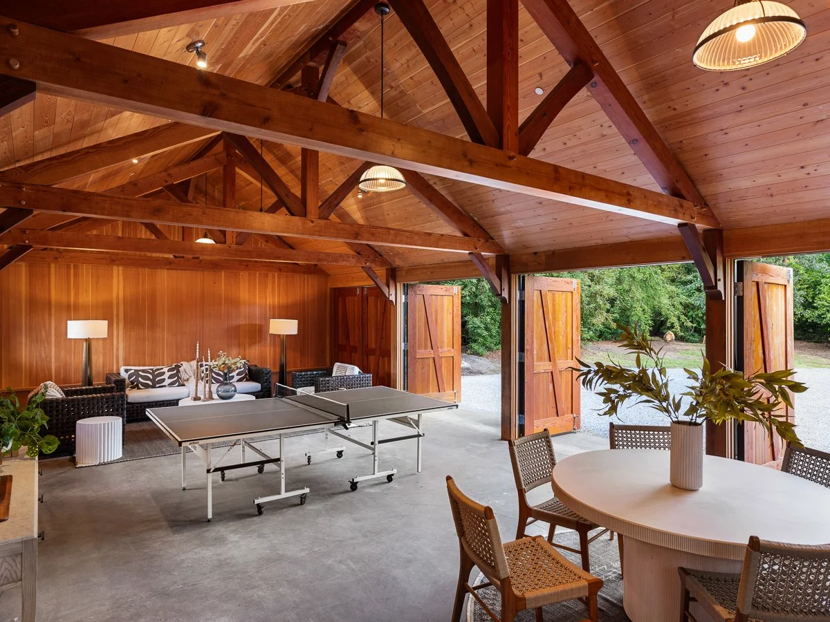 Interior of a wooden garage with a ping pong table, a round dining table with chairs, a sofa, lamps, and open garage doors showing outdoor greenery.