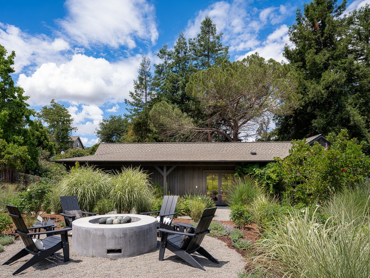 Outdoor backyard scene with a fire pit surrounded by four black chairs, lush green plants, and trees under a partly cloudy blue sky.