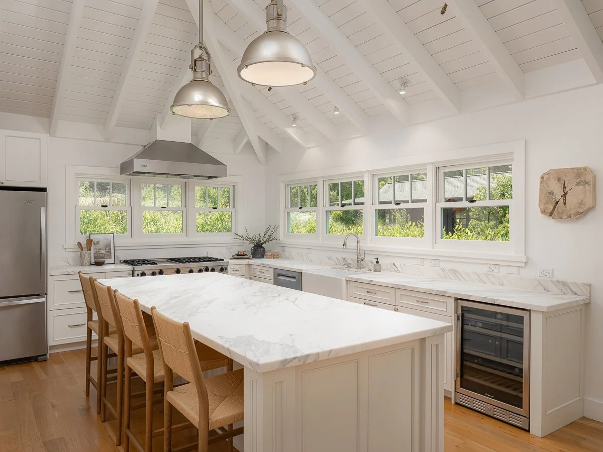 Bright white kitchen with high vaulted ceiling and exposed beams, stainless steel range hood, multiple windows, marble countertops, a kitchen island with four wooden chairs, small wine cooler, and modern fixtures.