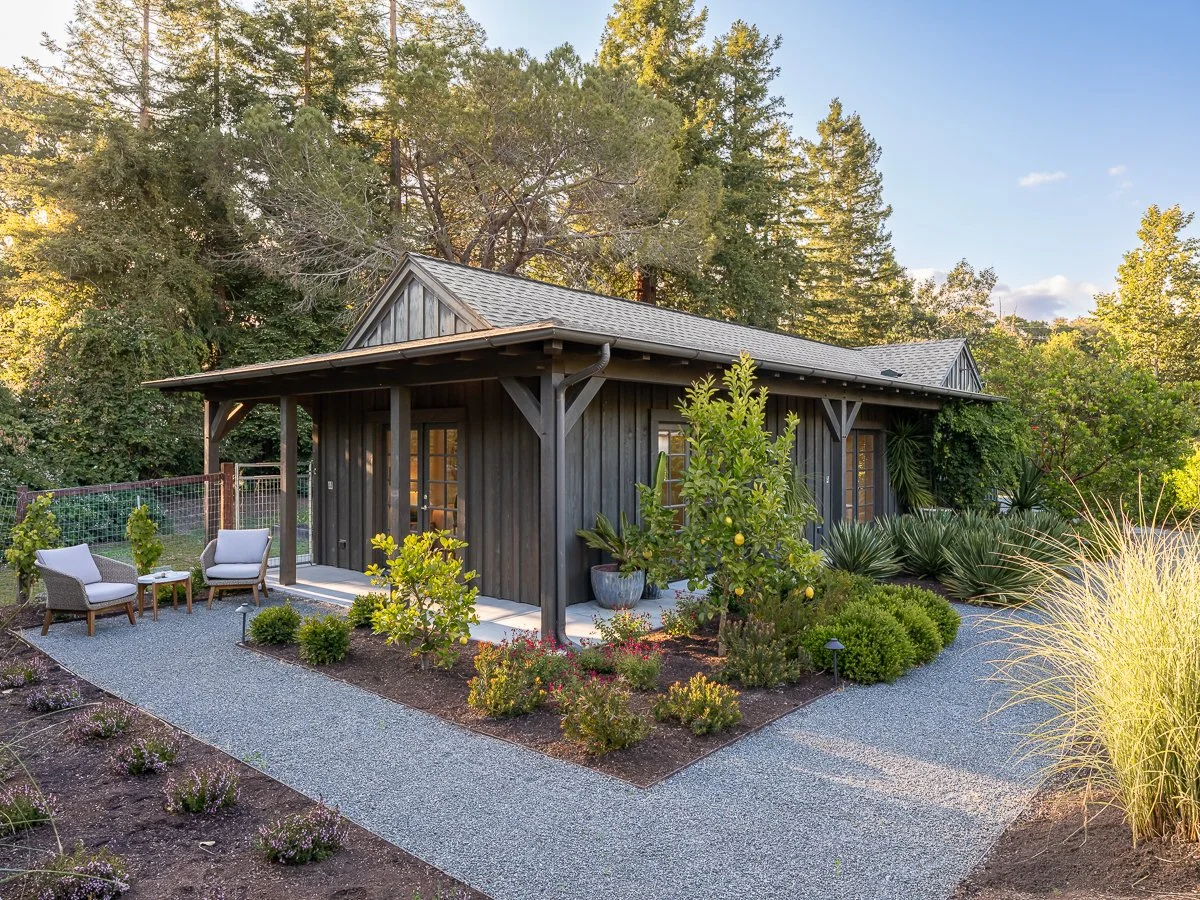 A small, dark wooden house with a porch, surrounded by a garden with bushes, trees, and gravel pathways on a sunny day.
