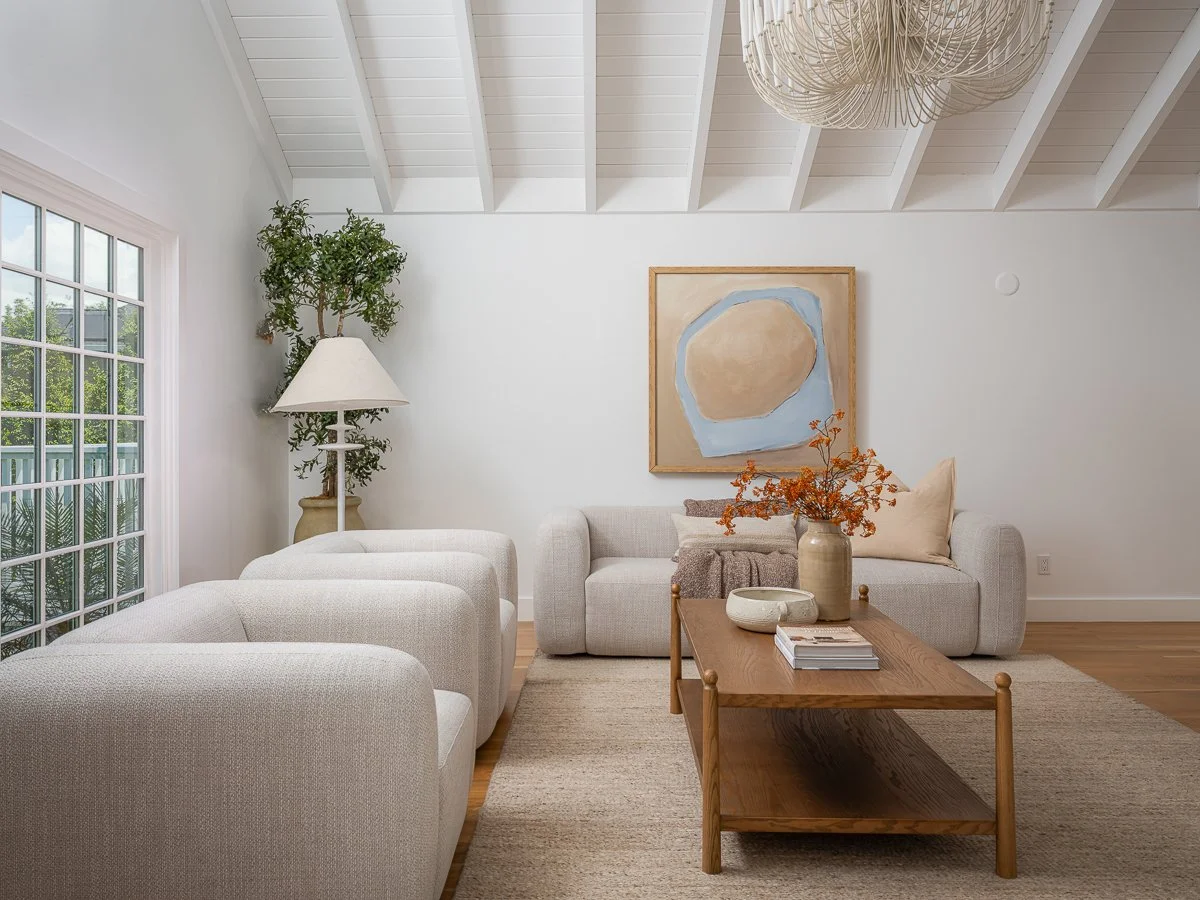 Living room with white walls and ceiling, beige sofa, three matching armchairs, a wooden coffee table with books and vases, a floor lamp, a potted plant, a piece of abstract wall art, and a large window with a view of greenery.