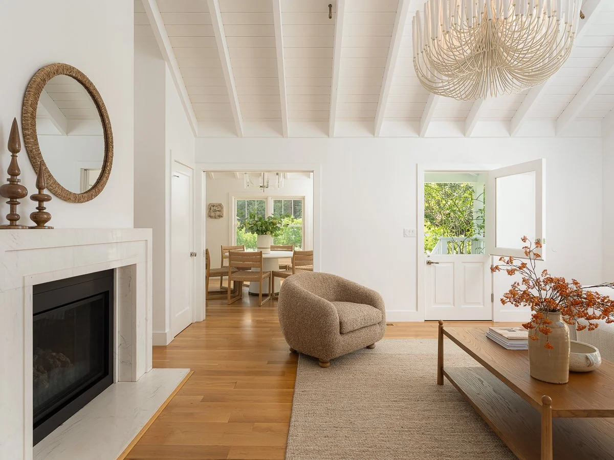 Bright living room with white walls, wooden flooring, a fireplace with a mirror above, a beige armchair, a wooden coffee table with a vase of orange flowers, and an open door leading to a balcony. In the background, a dining area with a round table a