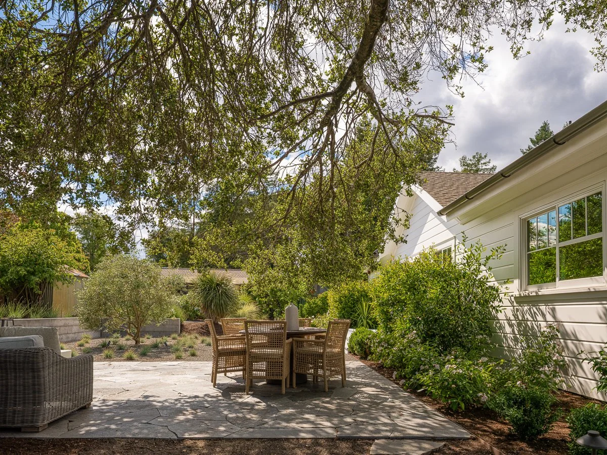 A backyard patio with outdoor wicker furniture, a round dining table, and surrounding lush green plants and trees next to a white house with a window. Overcast sky with scattered clouds.
