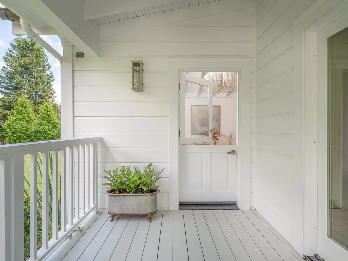 White porch with a potted fern, a door, and a window showing part of a living room with artwork and a hanging light fixture.