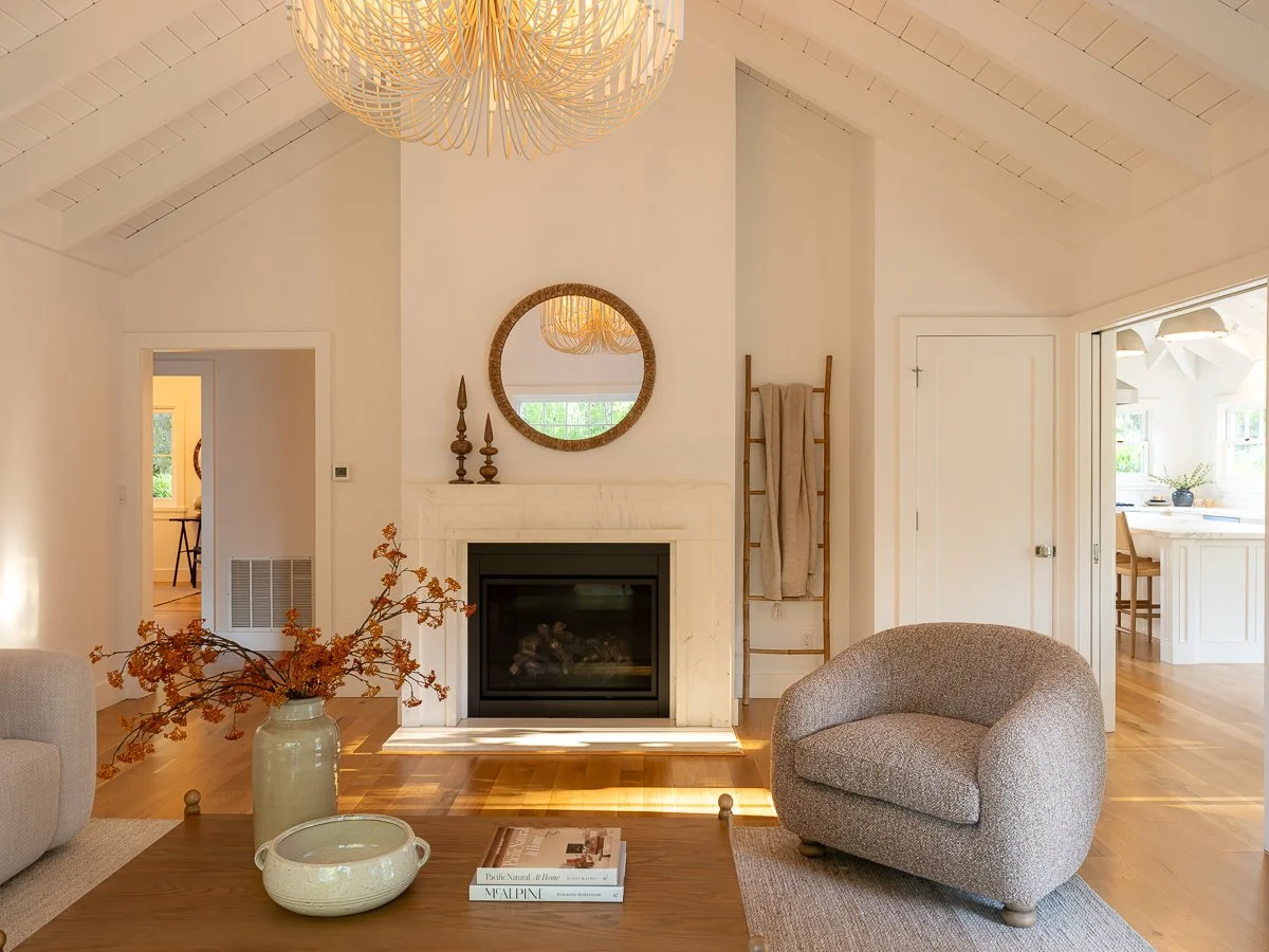 Living room with a beige armchair, a wooden coffee table with a large vase and a book, a fireplace with decorative candles on the mantel, a round mirror above the fireplace, a ladder with a towel, and natural light coming through windows.