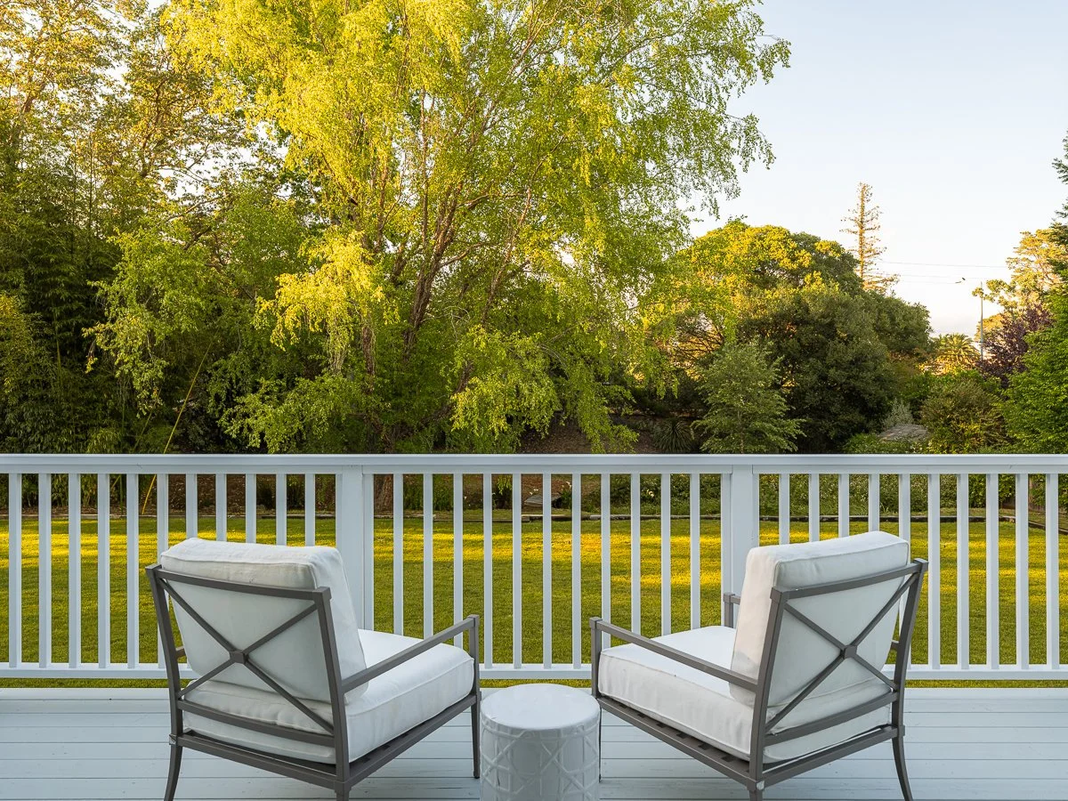 A view from a balcony with two white cushioned chairs and a small white side table, overlooking a grassy yard and lush green trees in the distance during daylight.