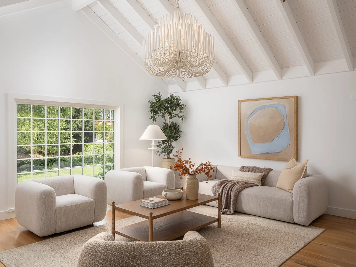 Bright living room with white walls, large window with greenery outside, and a high ceiling with white beams. Contains a white armchair, a sectional sofa with throw pillows and blanket, a wooden coffee table with decorative items, a tall potted plant