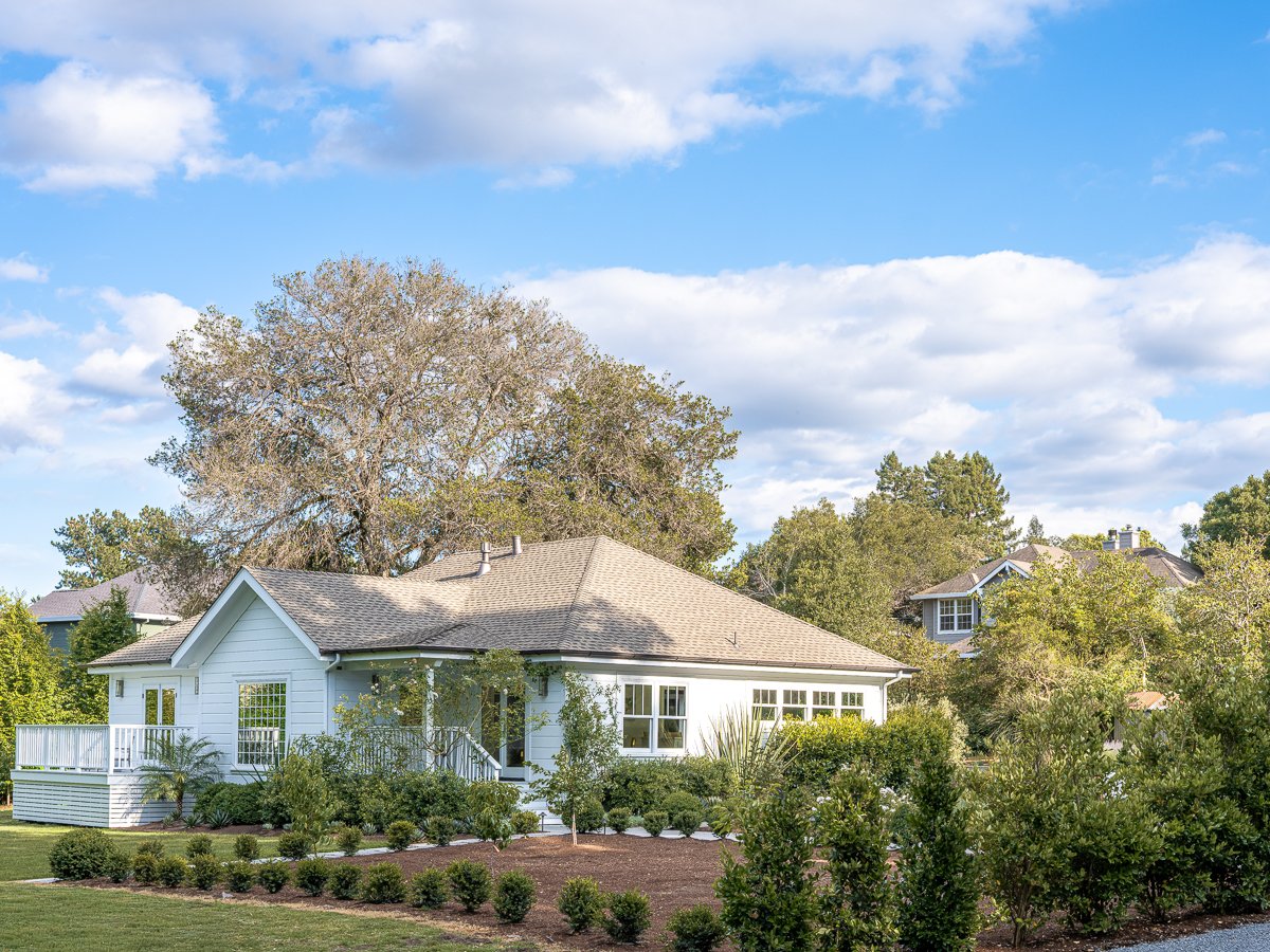 A white house with a gray roof surrounded by green trees and shrubs under a partly cloudy sky.