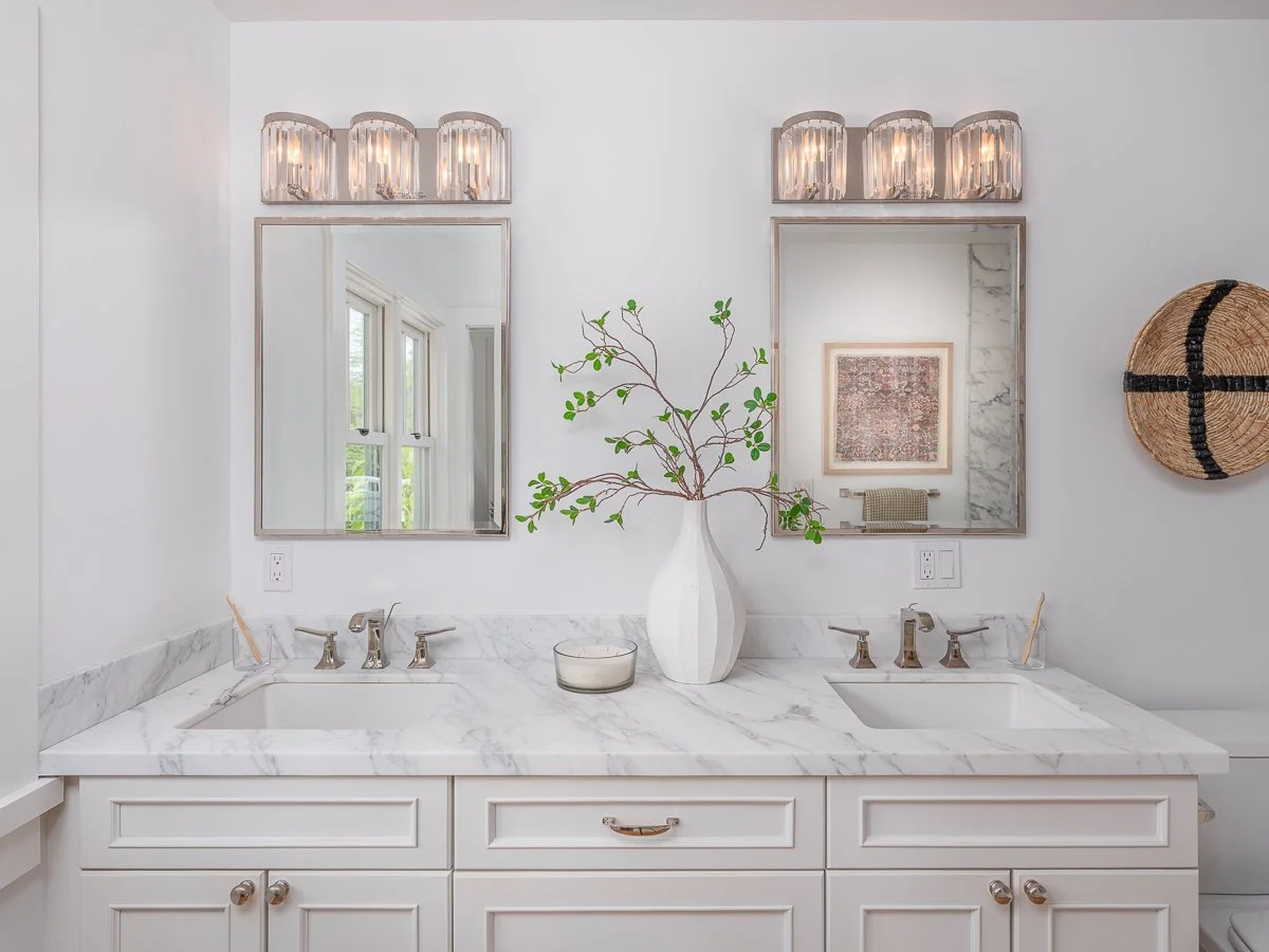 A modern bathroom vanity with a marble countertop, dual sinks, and two mirrors. A white vase with green branches is centered on the counter. There are wall-mounted light fixtures above each mirror, and a round woven wall decor piece on the right.