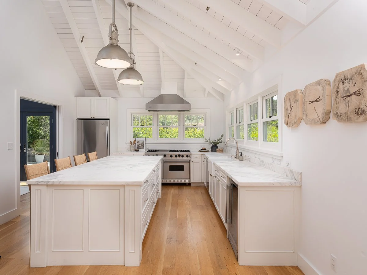 Bright white kitchen with vaulted ceiling, large island, stainless steel appliances, and multiple windows, featuring wooden flooring and light-colored cabinetry.
