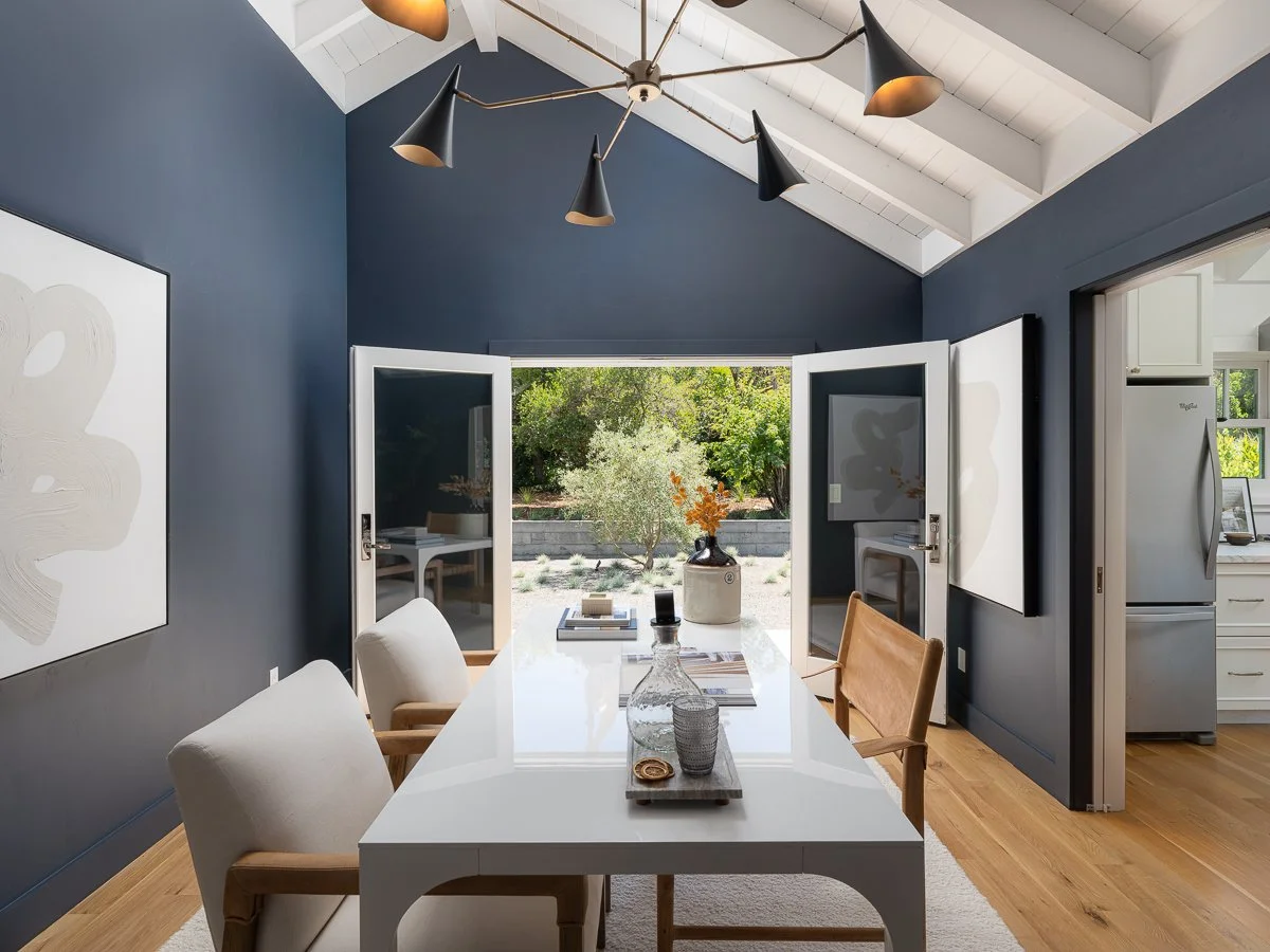 A dining room with a white table, four chairs, dark blue walls, and double doors open to a garden with trees and plants.