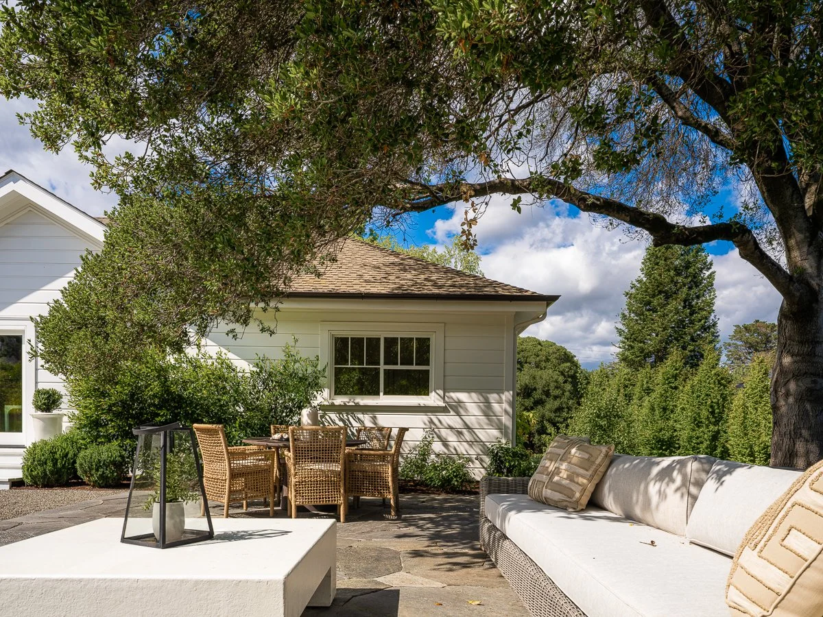 Outdoor patio area with wicker dining set, white sofa with beige cushions, tree providing shade, and greenery in the background.