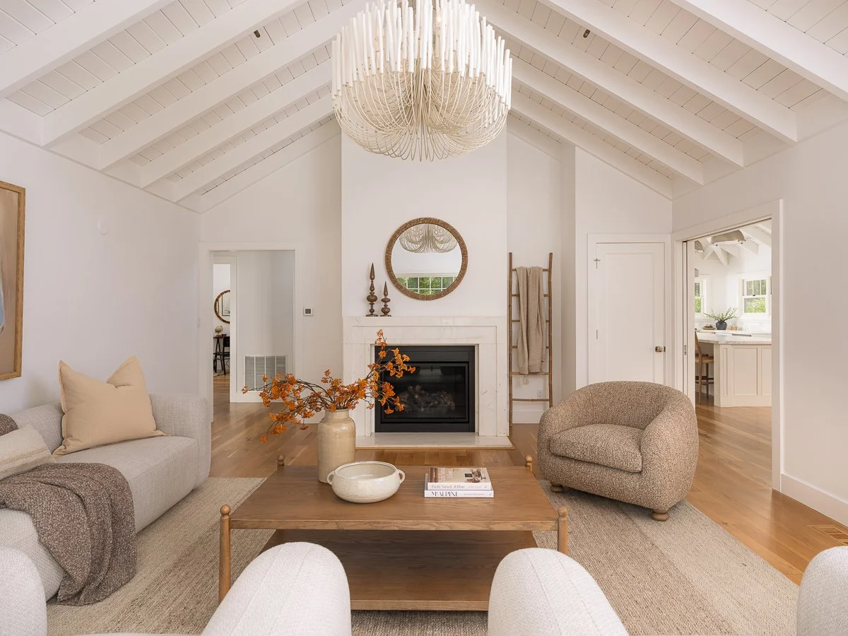 Living room with white walls and vaulted ceiling, beige sofa with pillows, round beige armchair, wooden coffee table with vases and books, fireplace with mirror above, wooden ladder with blanket, and chandelier hanging from ceiling.