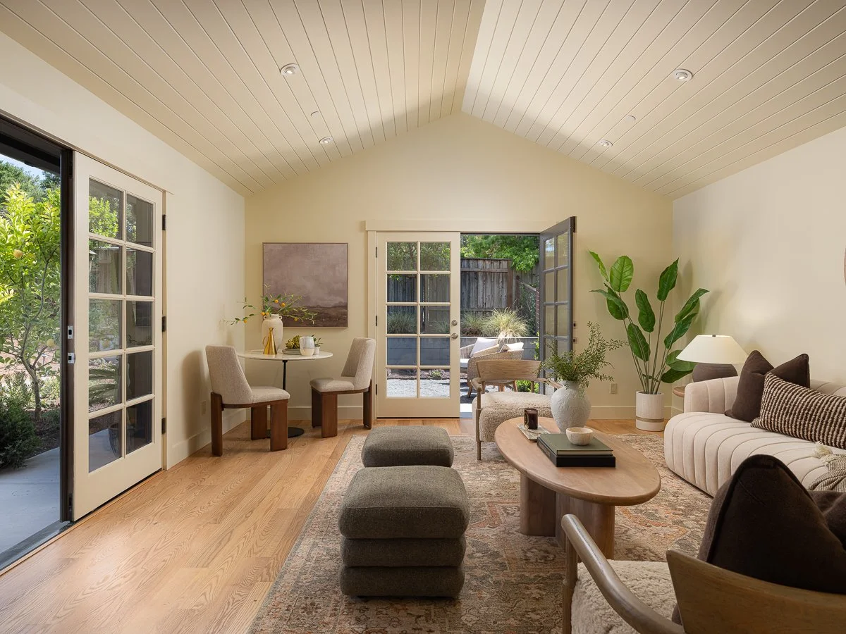 Living room with light wood flooring, beige walls, large glass door leading to outdoor patio, and comfortable beige sofa with brown pillows. There are two gray ottomans, a wooden coffee table with books and decor, a round dining table with four chair