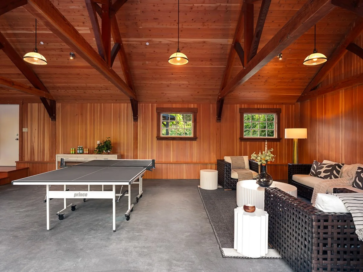 A spacious room with wood-paneled walls and ceiling, featuring a ping pong table on a concrete floor, and a sitting area with black wicker sofas, coffee tables, and a lamp, illuminated by hanging lights.