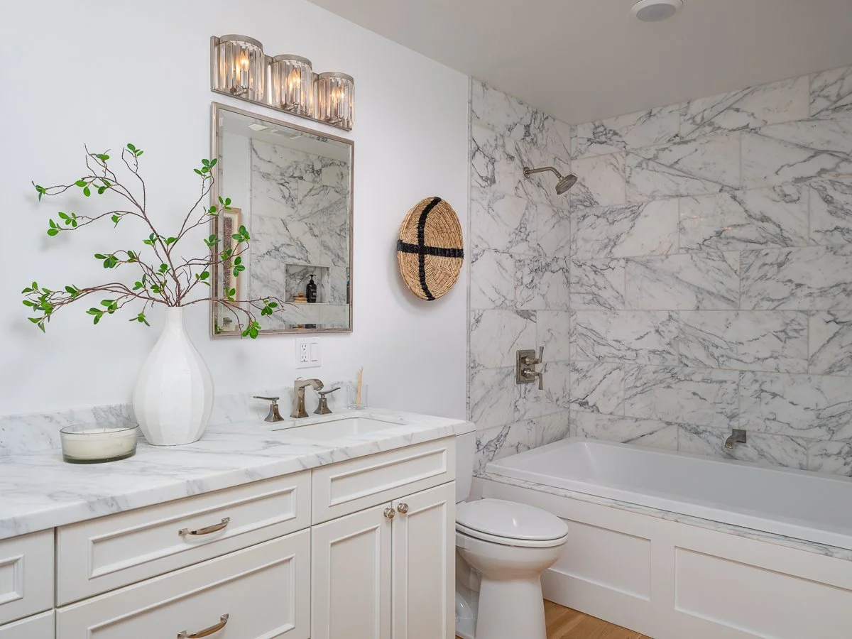 A bathroom with a marble vanity, a mirror, a vase with a branch, a soap dispenser, a toilet, a bathtub, and a shower with marble walls.