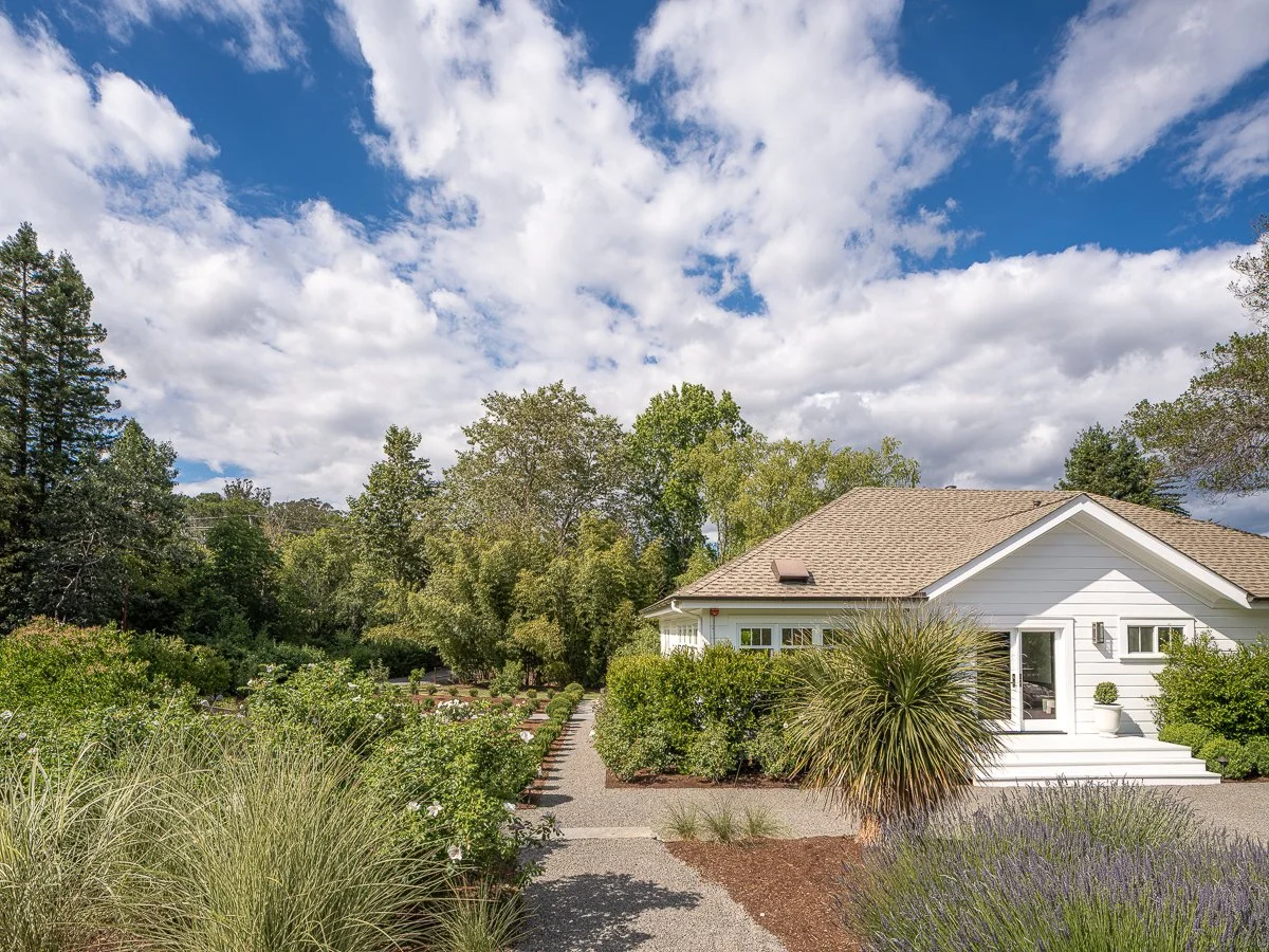 A white house with a brown roof and a sliding glass door on the front porch. The front yard has various bushes, plants, and trees with a gravel pathway leading to the house. The sky is partly cloudy with patches of blue.