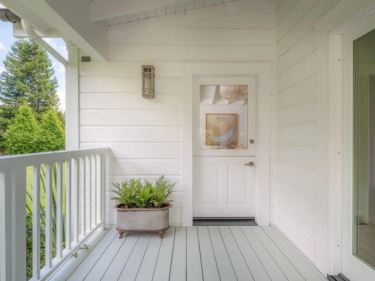 White porch with railing, potted fern plant, and white door with window, exterior of a house with trees and blue sky in the background.