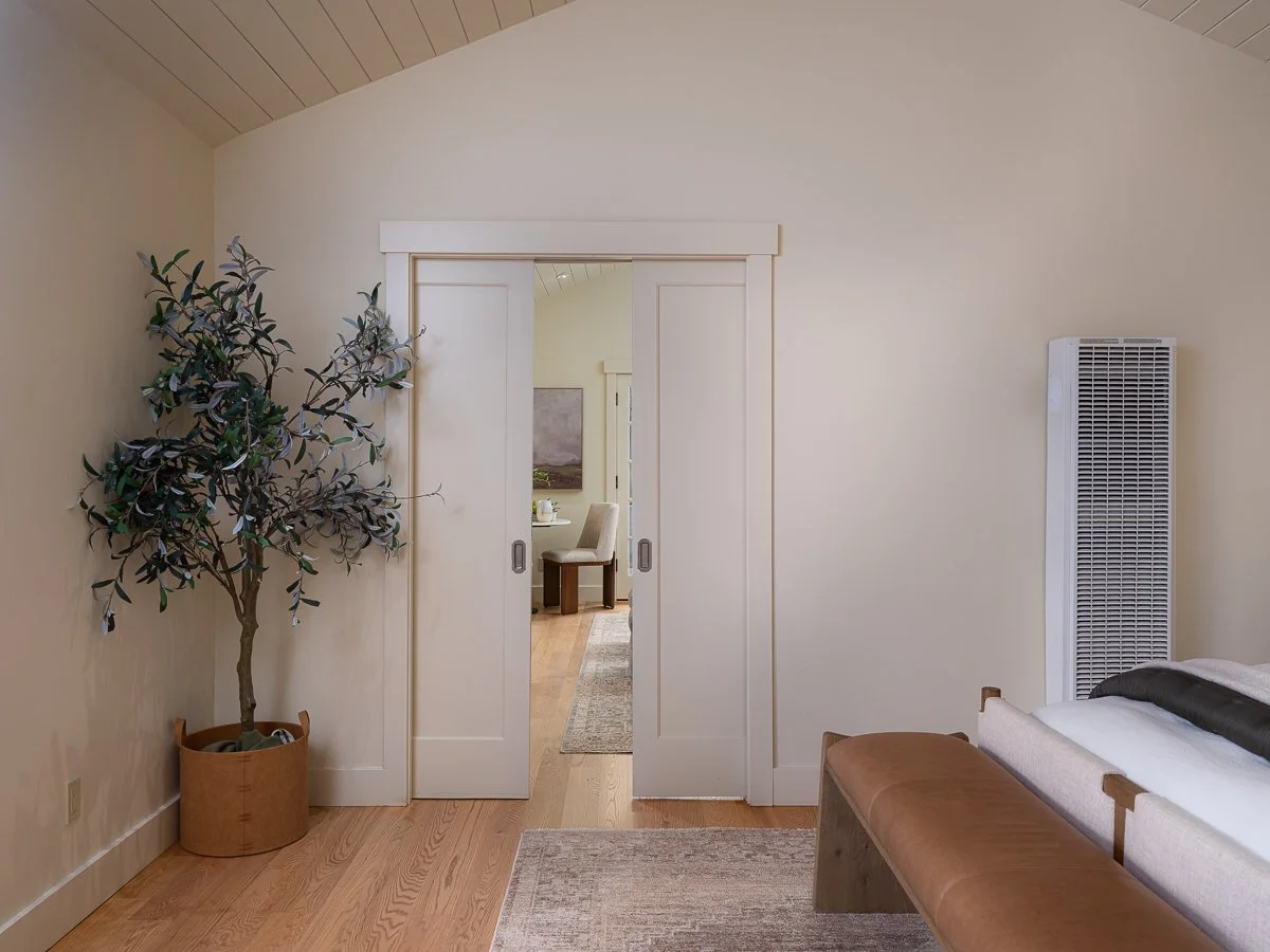 A living room with a potted plant on the left, a white closet door with sliding door panels, a beige bench, and a heater on the right. Through the open sliding doors, a room with a table and chairs is visible.
