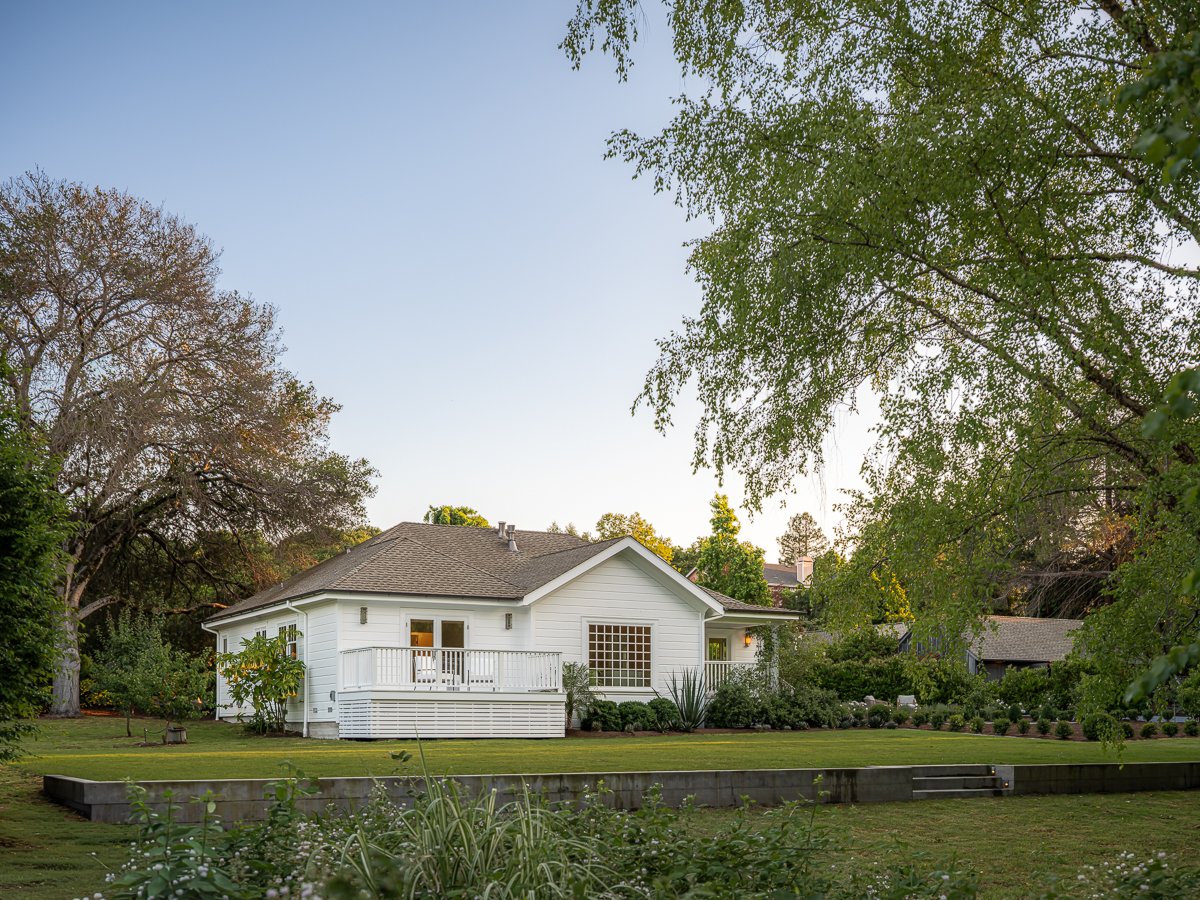 A white house with a porch in a lush green yard surrounded by trees, under a clear blue sky during daytime.
